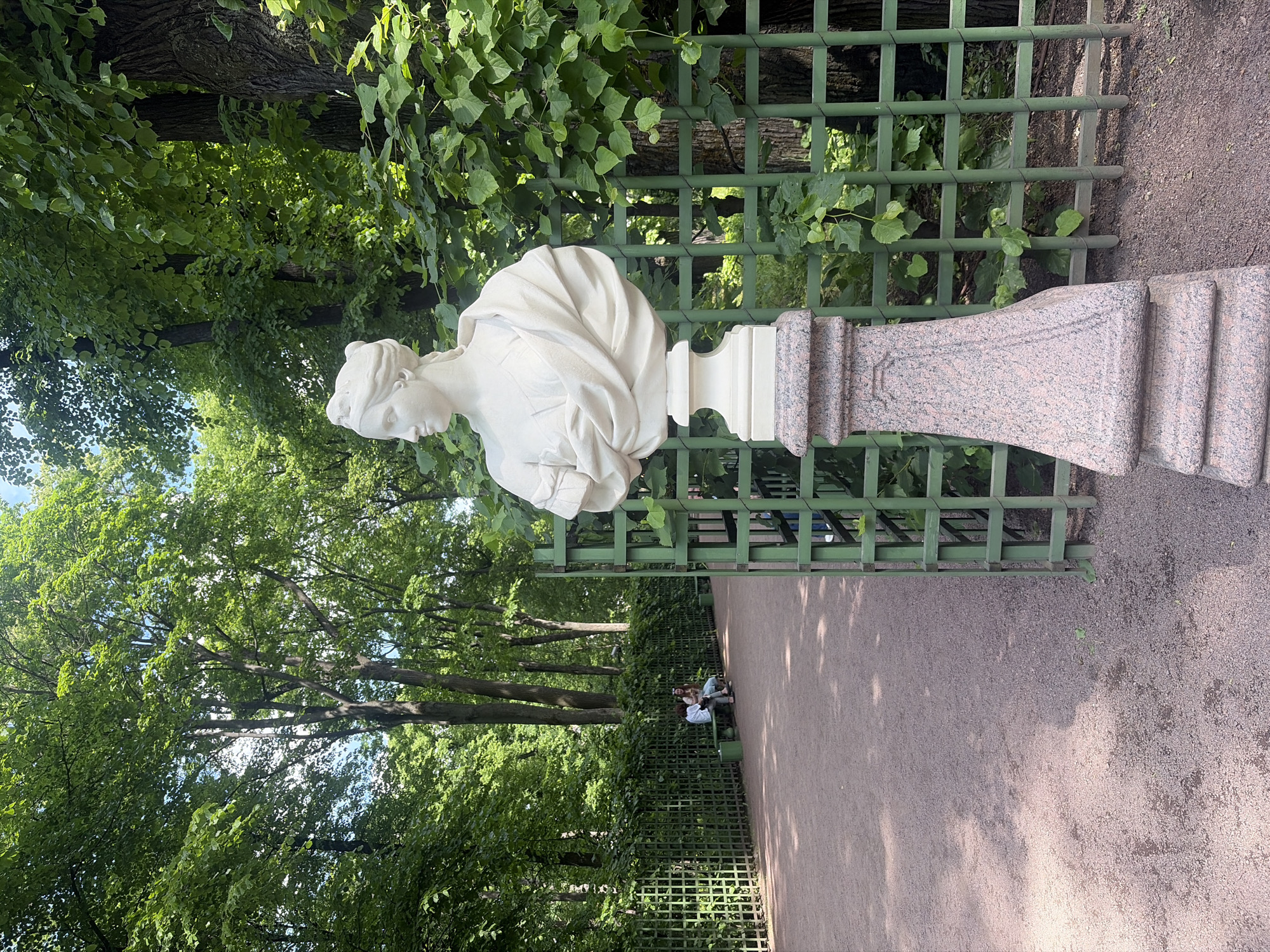 This is an outdoor photograph of a garden in Saint Petersburg, Russia, likely taken in the Summer Garden due to the ornate green lattice fencing and the presence of classical statuary. In the foreground, a white marble bust of a woman is displayed on a tiered stone pedestal, which is adorned with a granite base. The bust is positioned to the right of the frame, behind the green lattice fence. The fence extends diagonally across the image, creating a sense of depth and separating the foreground from the background.
The background features a dirt pathway that curves to the left, lined with the green lattice fence and lush, green trees. Sunlight filters through the leaves, creating dappled shadows on the ground. In the middle ground, two individuals are seated on a bench along the pathway, facing away from the camera. They appear to be engaged in conversation. The overall atmosphere is serene and picturesque, typical of a well-maintained historical park. The vegetation is dense and verdant, indicating a warm season. The sky is visible through the tree canopy, suggesting a bright, clear day.