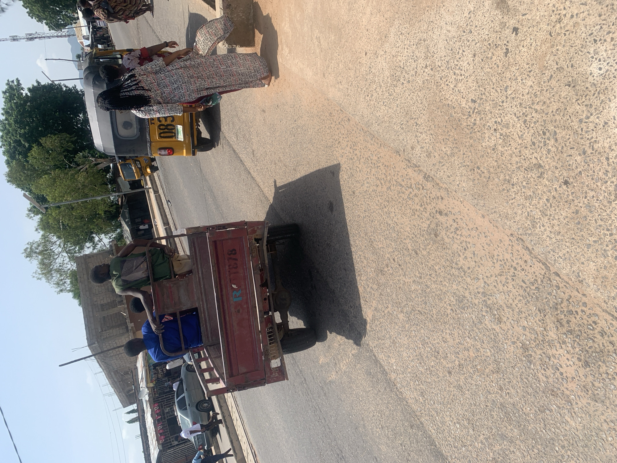 The image shows a street scene in Jalingo, Nigeria, under what appears to be bright daylight with a clear sky. A yellow auto-rickshaw, also known as a "Keke Napep," is prominently featured, with a number plate reading "C E 680." Two women are visible near the auto-rickshaw. One woman, wearing a patterned dress, is facing away from the camera. Another woman, with long dark hair, is also near the auto-rickshaw.
Behind the auto-rickshaw, a red flatbed trailer is being pulled. This trailer is occupied by three young men. The man at the front of the trailer is wearing a green t-shirt and is seated, leaning forward. The man behind him wears a blue shirt and is also seated. A third person is visible in the trailer, further back. The trailer has text on its side, including what appears to be "R 1878" in blue lettering.
The scene is set on a paved road with a rough texture. The background shows buildings, some appearing to be under construction, and lush green trees. There are also other vehicles, including a silver car and some smaller structures that might be stalls or shops, lining the street. The overall impression is of a busy, everyday street scene in a Nigerian town. The lighting suggests it is daytime, likely mid-morning or afternoon, with the sun casting distinct shadows.
