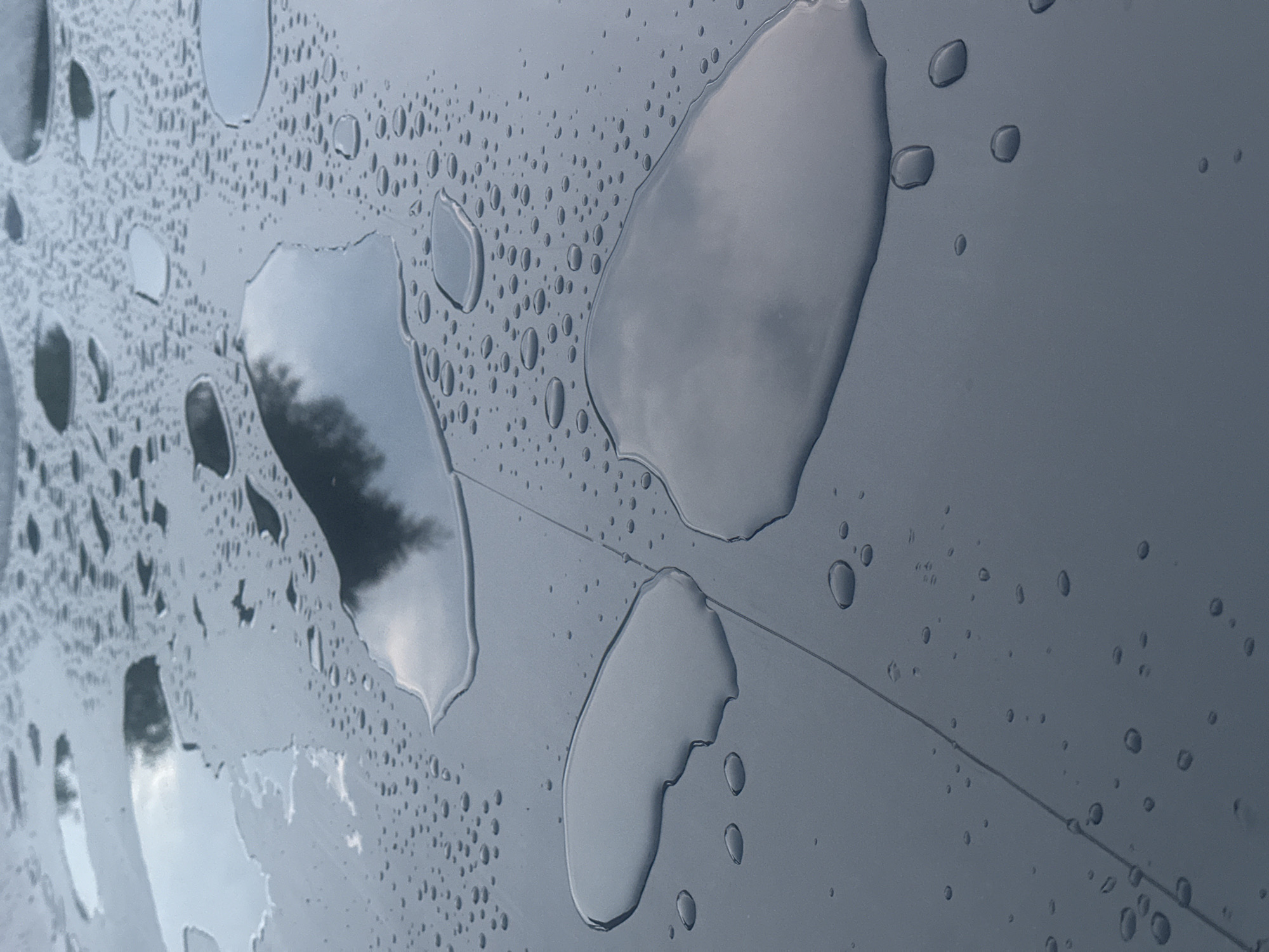 The image shows a close-up, high-angle view of a dark, smooth, flat surface covered in water droplets and several larger puddles. The surface appears to be outdoors, with reflections of a cloudy sky and possibly trees visible in the larger puddles. There are no people, animals, or specific landmarks visible that would indicate a location within the city of Lapės, Lithuania, or any other specific urban setting. The scene is purely naturalistic, focusing on the abstract patterns created by the water on the surface. The surface itself is segmented by a subtle, straight line running diagonally through the image, suggesting it might be a tiled or paneled structure.