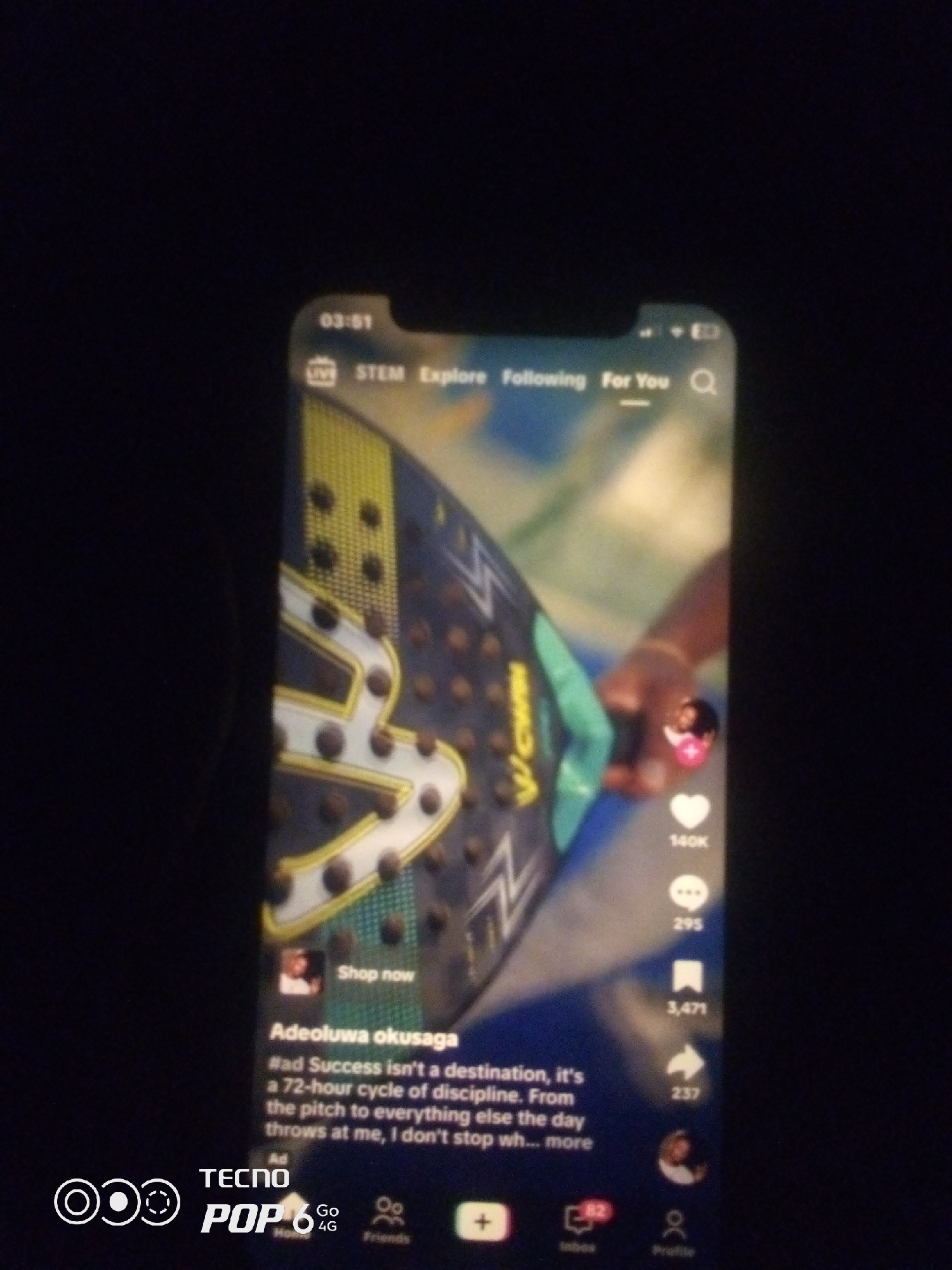 The image shows a smartphone screen displaying a social media application, likely TikTok, with a video playing. The video appears to feature a hand holding a tennis racket with a distinctive design. The screen displays standard app interface elements such as time, navigation tabs ("STEM", "Explore", "Following", "For You"), user profile icons, like counts ("140K", "295", "3,471", "237"), and comment/share icons. There's also text overlay describing the video content, including a hashtag "#ad Success isn't a destination, it's a 72-hour cycle of discipline. From the pitch to everything else the day throws at me, I don't stop wh... more". The bottom of the screen shows app icons for home, inbox, and profile, along with branding for "TECNO POP 6 4G". The image is taken in a dark environment, with the phone screen being the primary light source. There is no visible indication of the location being Howayi, Nigeria, or any other specific city.