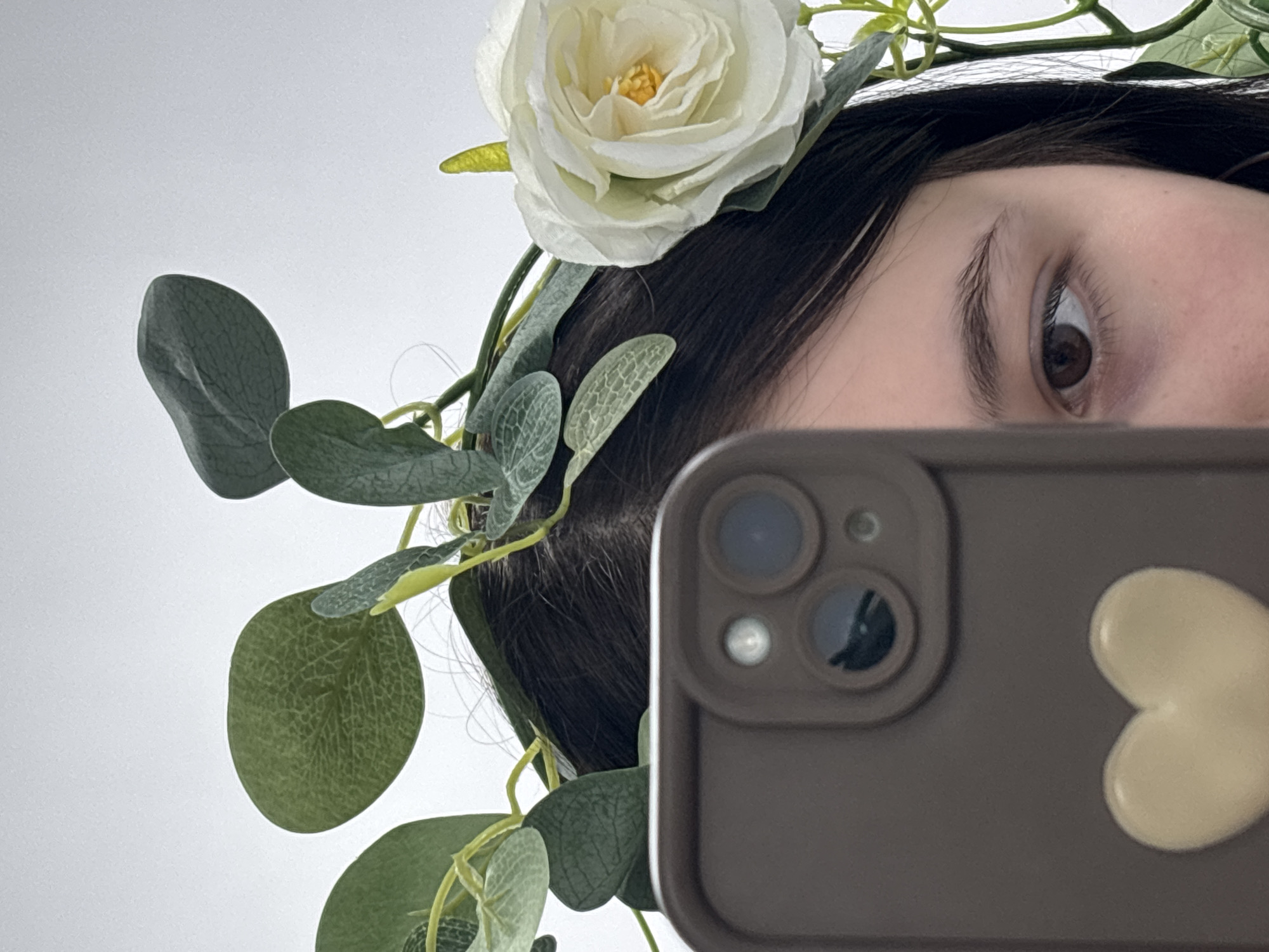 A close-up selfie, taken in Фоки, Russia, shows a person's partially visible face framed by a floral headpiece against a plain, light-colored background. The right side of the person's face is visible, including a dark brown eye, an arched eyebrow, and strands of dark hair. A prominent white rose with a yellow center sits atop the person's head, surrounded by numerous green, ovate, eucalyptus-like leaves and delicate vines, forming a garland.
In the lower right foreground, a dark brown or gray smartphone is held up, its camera array featuring multiple circular lenses and a flash visible. A light-colored, heart-shaped sticker is attached to the back of the phone, next to the camera module. The person's eye is directed towards the smartphone's lens, indicating they are taking the photo themselves. The setting appears to be indoors. No text is visible in the image.