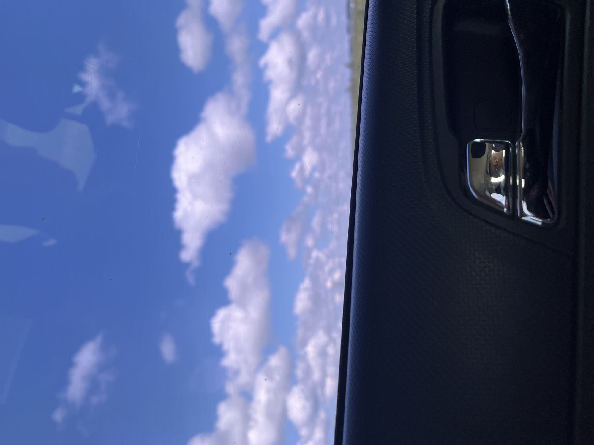 The image is taken from inside a vehicle, looking out through the window at a bright blue sky with scattered white cumulus clouds. A portion of the vehicle's interior is visible in the foreground, including a black textured armrest and a chrome-plated door handle. The reflection on the chrome handle shows a person's face. The background appears to be a slightly blurred landscape with a faint horizon line. The image was captured in the city of Марьевка, Russia.