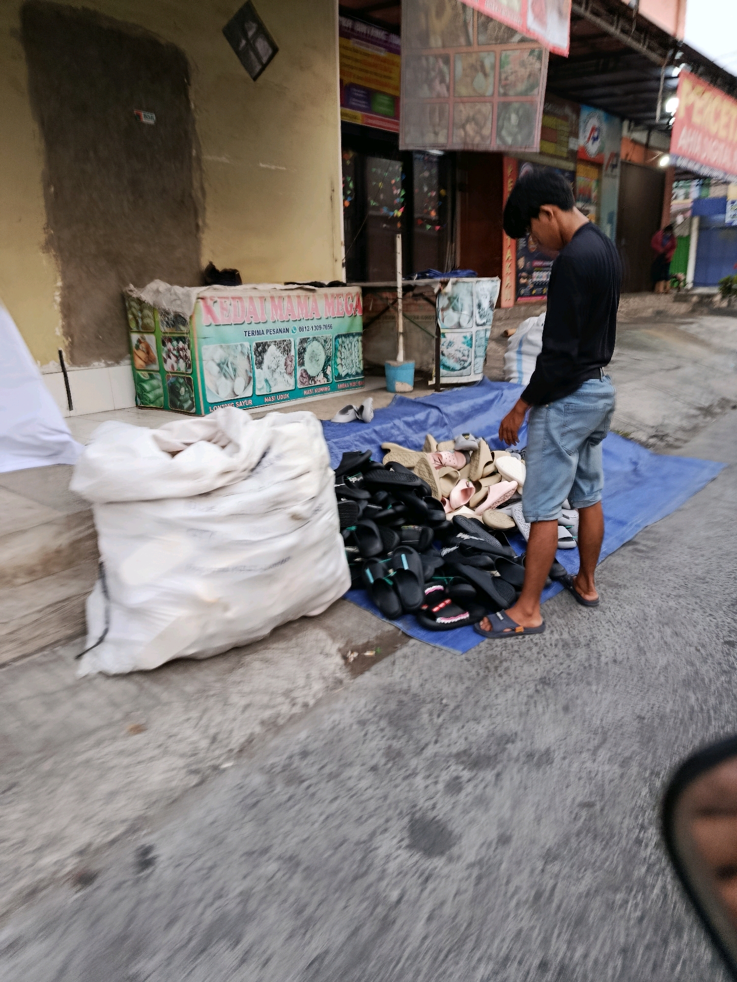The image depicts an outdoor scene on a street in Bekasi, Indonesia. In the foreground, a man wearing a black long-sleeved shirt and denim shorts is standing next to a pile of footwear, primarily flip-flops and sandals, laid out on a blue tarp. The man appears to be interacting with the footwear, possibly organizing or displaying it. To the left of the man is a large white sack, likely containing goods. In the background, there is a building with signage that appears to be a shop or food stall, with text that reads "KEDAI MAMA MEGA". The wall of the building is a pale yellow with a textured section that looks like it might be under repair or has been plastered. The overall environment suggests a commercial street setting.
