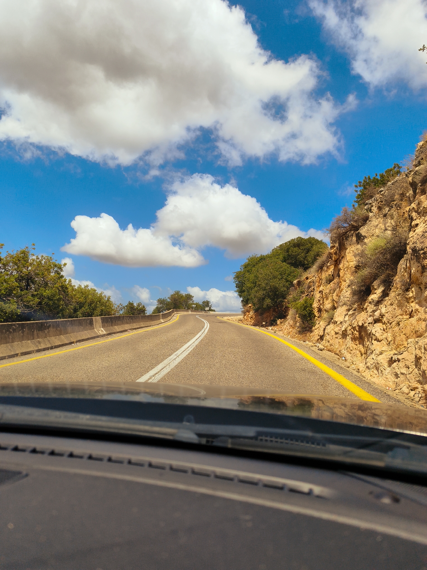 The image depicts a view from the interior of a vehicle traveling on a paved road. The road curves to the right, bordered by a concrete barrier on the left and a rocky, vegetated hillside on the right. The sky is blue with scattered white clouds. The location is identified as a road in the city of Mate Asher Regional Council, Israel. No people or other subjects are visible.