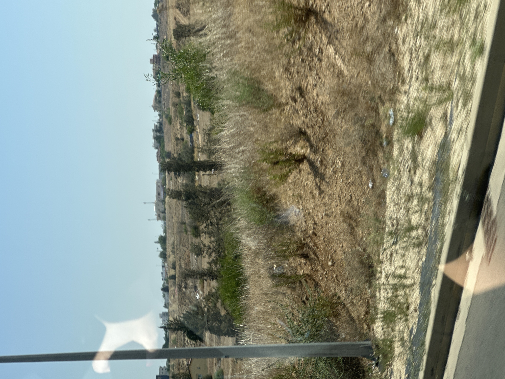 The image depicts a dry, arid landscape with sparse vegetation and a few scattered buildings in the distance. A gray pole is visible on the left side of the frame, possibly a lamppost or utility pole. The ground is covered in dry grass, dirt, and some scattered debris. The sky is clear and blue. The overall impression is of a rural or semi-developed area with a dry climate.
