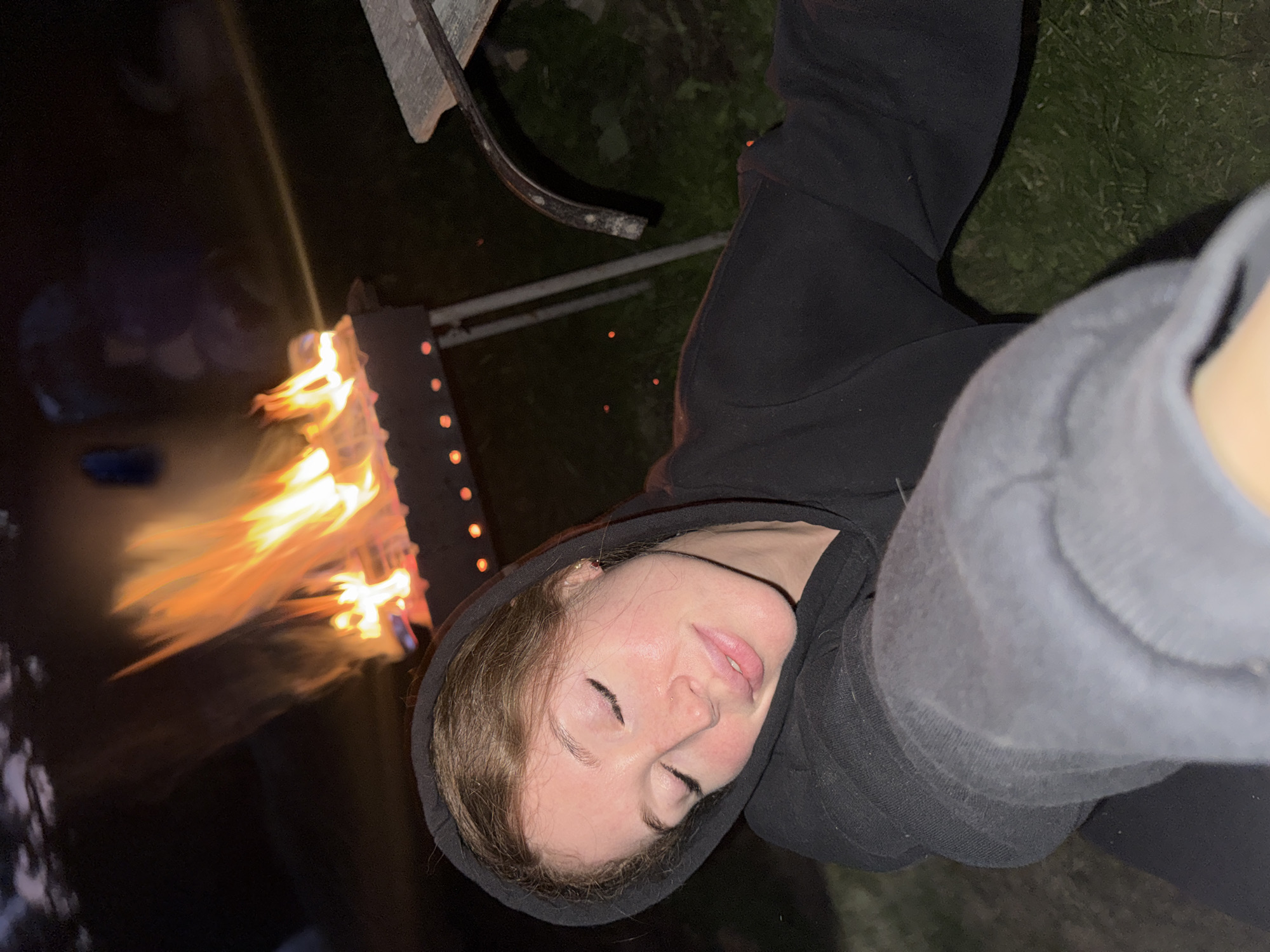 A young woman with reddish-brown hair and closed eyes is captured in a close-up selfie. She is wearing a black hoodie, with the hood pulled up over her head. Her eyes are shut, and her face has a serene expression, with her head tilted slightly. In the background, a bonfire burns brightly in a metal grill, casting an orange glow on the scene. The environment appears to be outdoors at night, with grass visible on the ground and hints of other people and objects in the dark periphery. The location is identified as Поварёнки, Russia.