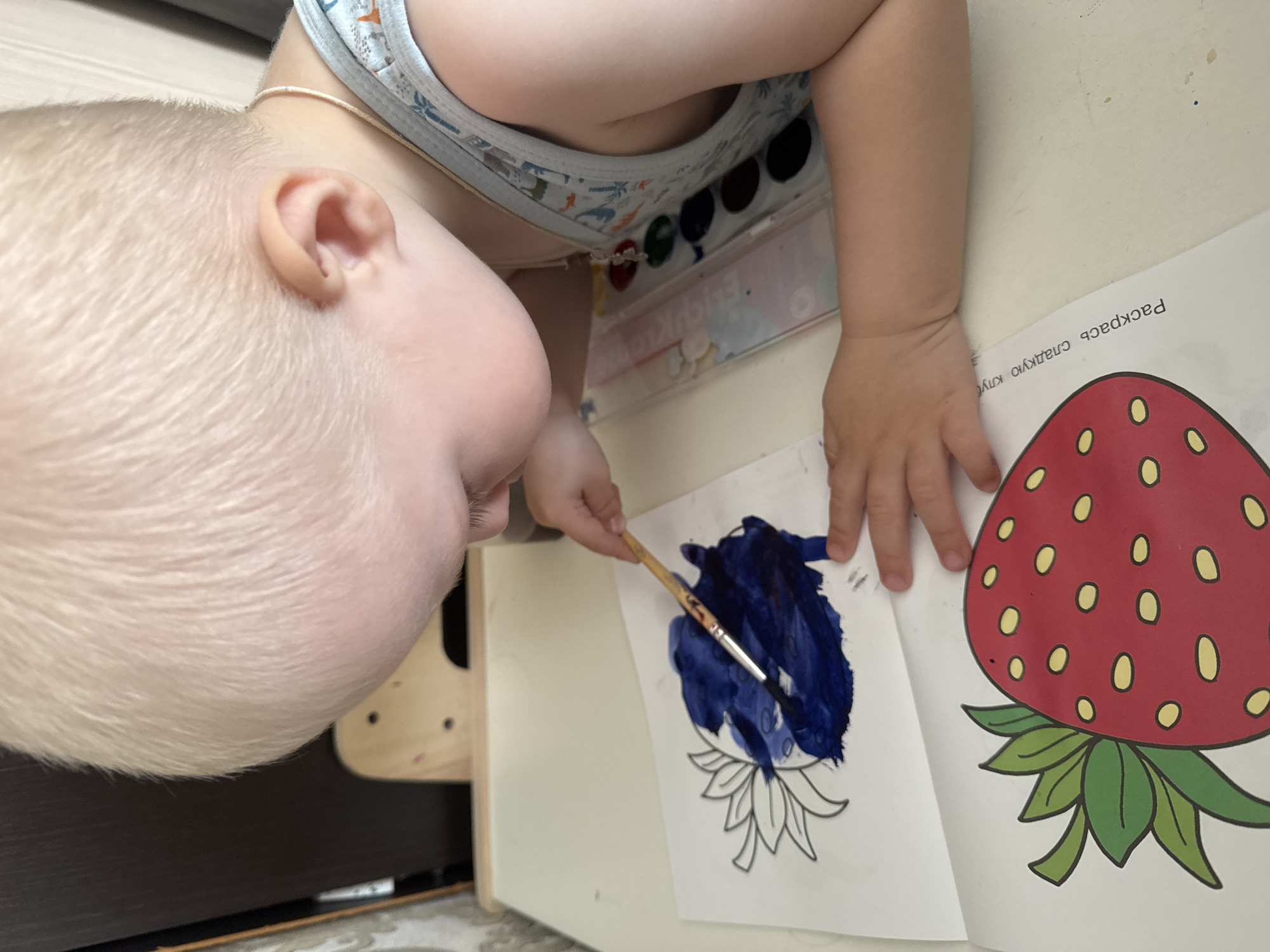 A young child with blond hair is painting a picture of a flower with dark blue paint on a white sheet of paper. The child is wearing a light-colored diaper with a pattern. Next to the flower drawing is a drawing of a strawberry that has already been colored in. There is a palette of paints visible near the child's arm. Text in Russian is present on the paper with the strawberry image. The setting appears to be indoors, on a light-colored table. The photograph was likely taken in the city of Chaikovsky, Russia, due to the presence of Russian text.