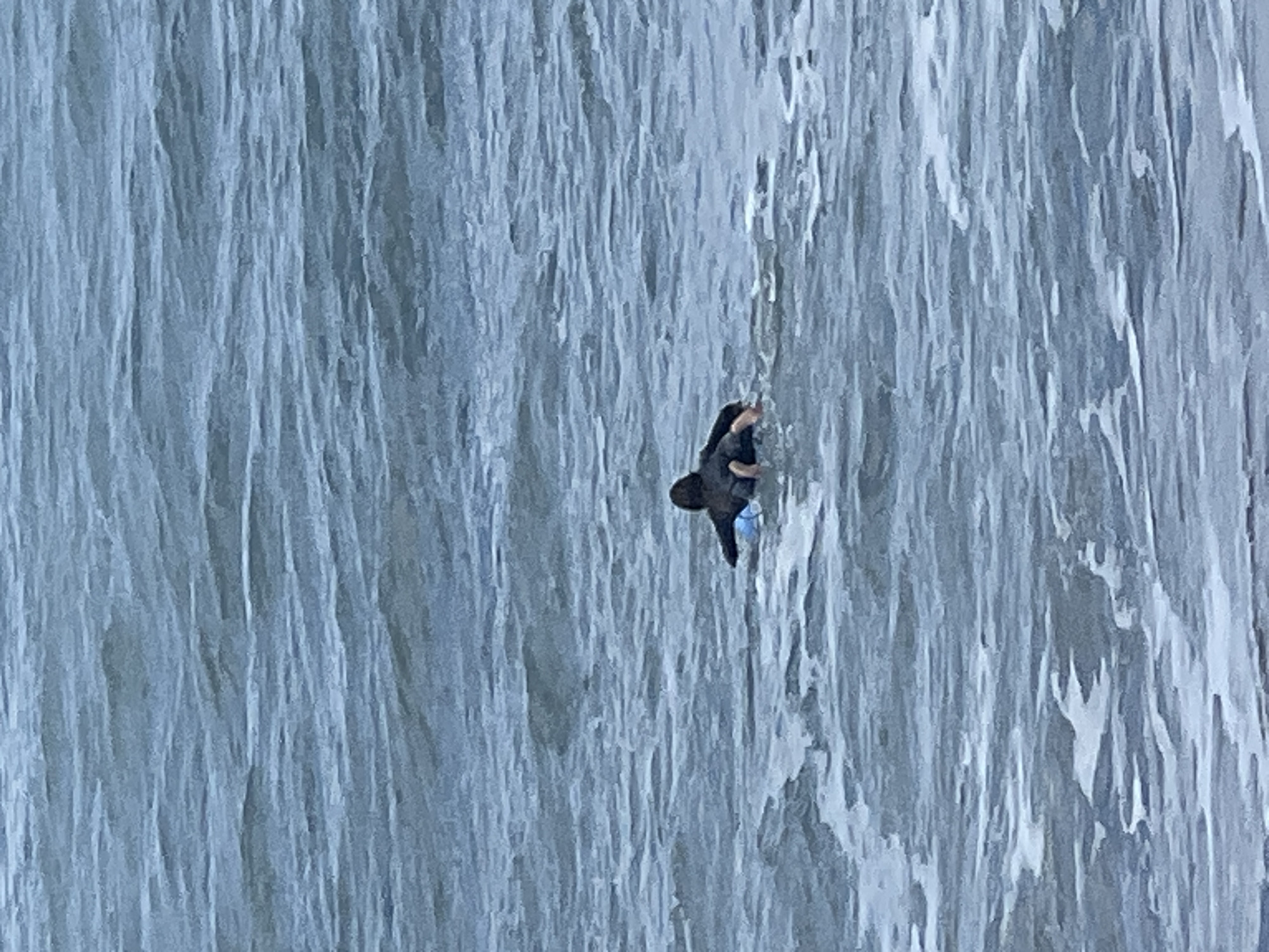 A lone individual is depicted engaged in an activity resembling climbing or rappelling on a colossal vertical wall. The wall, which dominates the frame, exhibits strong vertical striations in various shades of blue-grey and white. This texture closely mimics that of a massive frozen waterfall or an extensive ice formation, with many vertical grooves and ridged surfaces.
Given the provided location context of Las Palmas, Spain, it is highly probable that this is an artificial structure, such as a concrete dam wall, a purpose-built ice climbing simulator, or a similarly textured man-made facade designed to resemble natural ice or rock, rather than natural ice itself.
The person is dressed in dark attire, including what appears to be a jacket and trousers, and is equipped with a small backpack or climbing gear. Their posture suggests active movement, with limbs spread to maintain grip and balance on the textured surface. The immense scale of the structure is evident when comparing its size to the human figure. The scene is illuminated by daylight, but specific time of day or weather details are not discernible. No text is visible in the image.