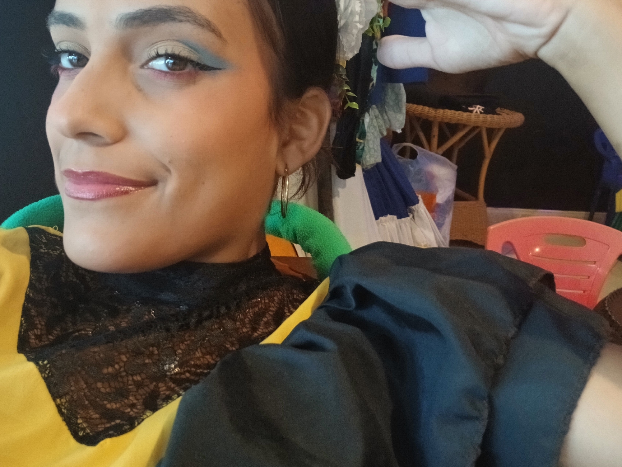 A woman is shown in a close-up, partially obscured by a dark blue fabric or garment draped over her shoulder and arm. She has brown eyes with distinctive eye makeup, including light eyeshadow and dark eyeliner, and glossy pink lipstick. She wears gold hoop earrings and a garment with a black lace collar over a yellow shirt. A green cushion is visible behind her neck. The background includes a black wall, a rattan table, miscellaneous clothing, and a pink plastic chair. There is no verifiable information in the image to confirm the location is Playa El Agua, Venezuela.