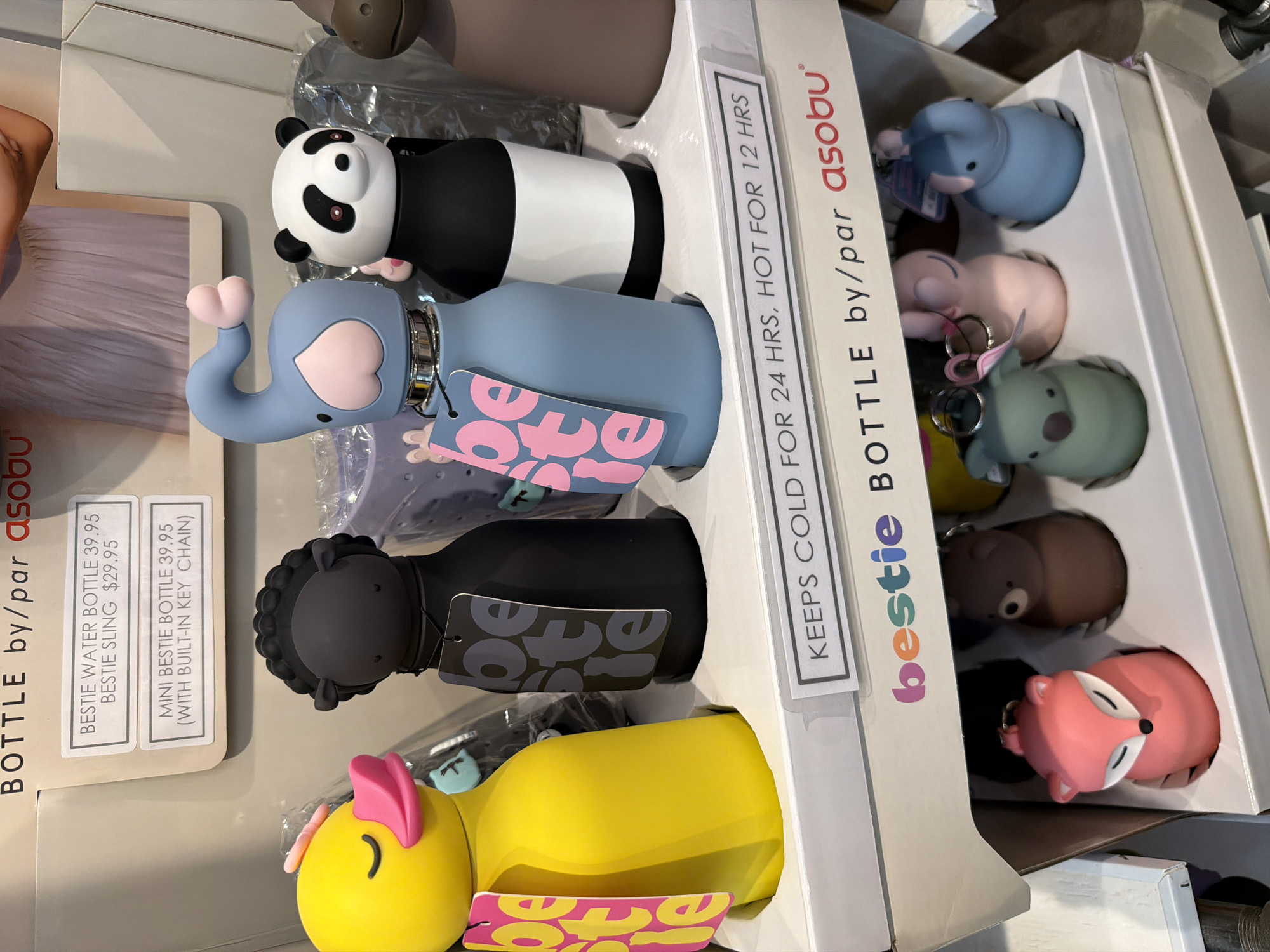 The image shows a display of "Bestie" brand water bottles and related accessories, likely in a retail store. The items are arranged on a white tiered display stand.
The top tier features several character-themed water bottles: a yellow duck with pink accents, a black sheep, a light blue elephant, a panda, and a brown bear. Each bottle has a tag attached. Above these bottles, on a white background, are two price labels. The top label reads "BESTIE WATER BOTTLE $39.95 BESTIE SLING $29.95". The bottom label reads "MINI BESTIE BOTTLE $39.95 (WITH BUILT-IN KEY CHAIN)". In the background, there is a blurred image of a person wearing a light purple dress.
The bottom tier of the display shows smaller, novelty keychain-like items, also shaped like animals. Visible are a pink fox, a brown bear, a green koala, a pink elephant, and a light blue elephant. These are also displayed in individual slots on a white surface.
A white banner across the middle of the display reads "KEEPS COLD FOR 24 HRS, HOT FOR 12 HRS". Below this, a larger "bestie" logo with colorful lettering is visible, followed by "BOTTLE by/par asobu".
The scene is brightly lit, and the focus is on the products. The environment suggests a point-of-sale display in a shop. The location of Niagara-on-the-Lake, Canada, is not evident from the visual information in the image itself.