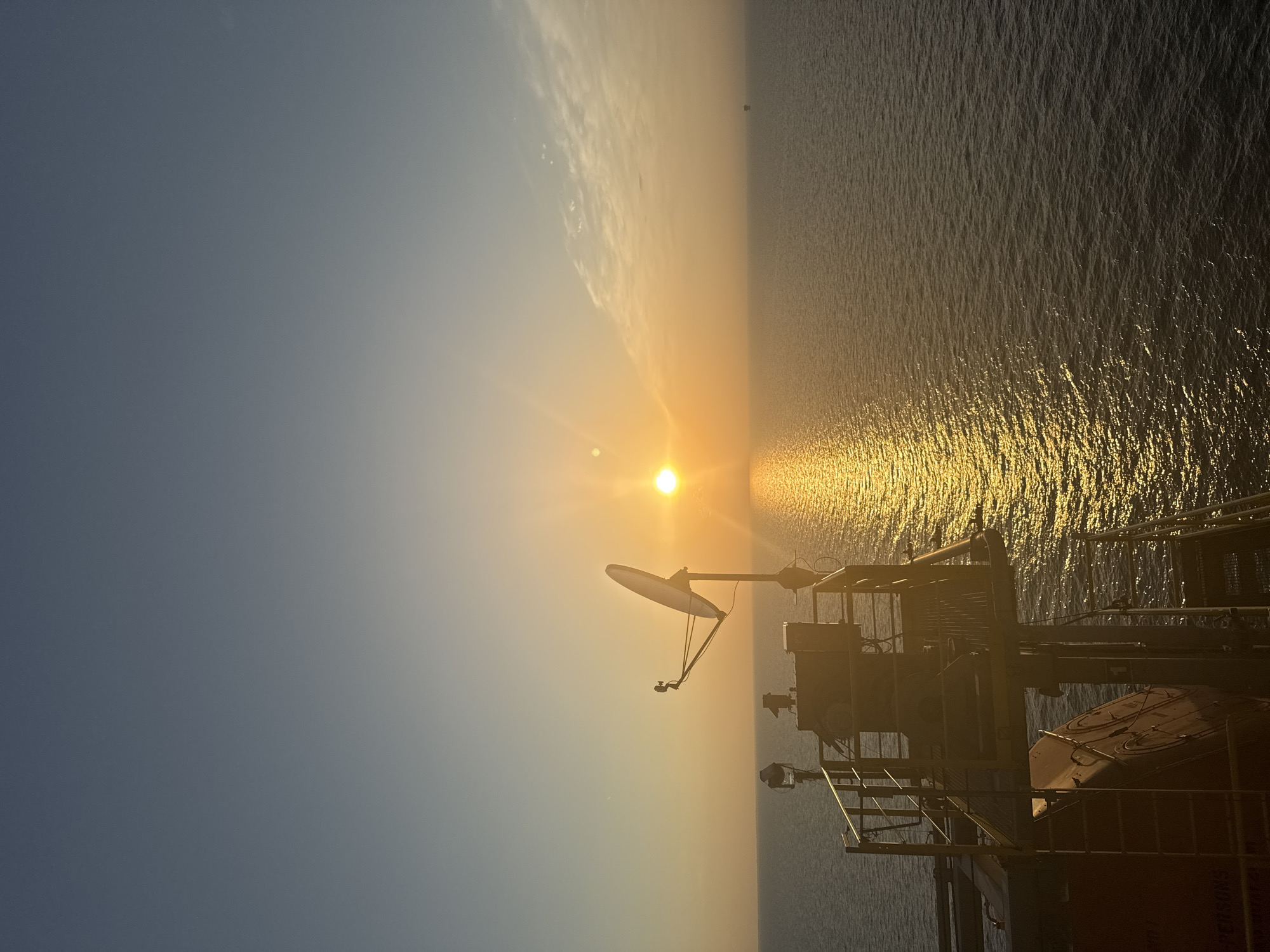 The image depicts an ocean scene at sunset. The sun, a bright orb, is low on the horizon, casting a golden path of light across the rippling water. A large satellite dish on a platform is prominent in the foreground, silhouetted against the warm sky. Several structures, including what appears to be a lifeboat, are also visible on the platform. In the distance, a small, dark structure can be seen on the water, possibly an oil rig or buoy. The sky transitions from a pale blue at the top to a warm orange and yellow near the horizon, with a few wispy clouds present. The overall impression is one of solitude and the vastness of the sea.