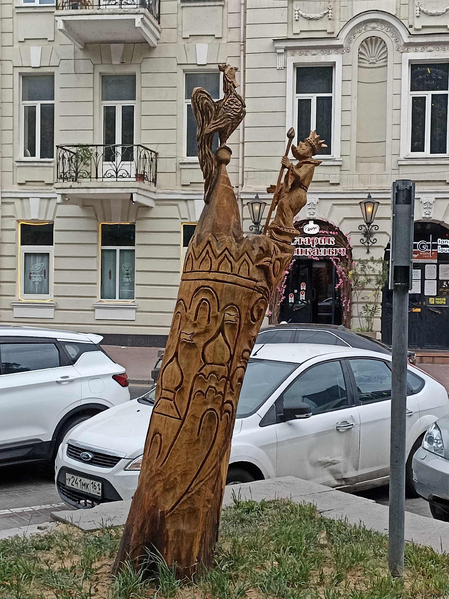 This is a medium shot, eye-level photograph of a wooden sculpture and the surrounding urban environment in Rostov-on-Don, Russia. The sculpture, made from a roughly carved tree trunk, depicts two figures. The top figure is a rooster, and below it is a figure wearing a crown and holding a staff, possibly a king or a royal figure. The trunk itself is intricately carved with various patterns.
In the background, a multi-story building with a light-colored facade is visible. It features several windows, balconies with wrought-iron railings, and decorative architectural elements. The building appears to be commercial, with some storefronts visible on the ground floor, including one with Cyrillic signage that reads "Старик Шкалик" (Old Man Shkalik). One of the storefront windows has a sign with "260" and another with "385" on it, likely prices or identification numbers.
In the foreground and midground, several white cars are parked along the street. One car in the foreground is a white Ford Focus, with the license plate "Y241MK 161". Another white SUV is parked to the left, and the rear of a gray car is visible to the right. The ground is a mix of pavement, grass, and a concrete curb. The lighting suggests an overcast day.