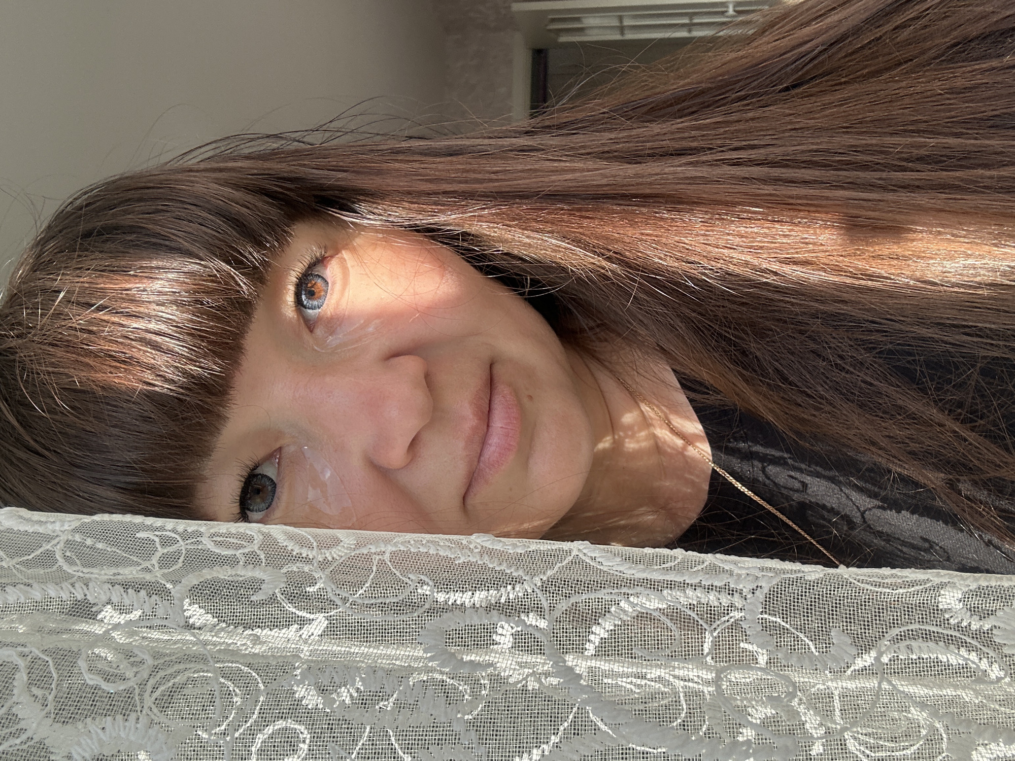 A woman with long brown hair and bangs is partially obscured by a white lace curtain on the left side of the frame. Sunlight illuminates her face, highlighting her eyes, which appear to have a gray-blue iris. A thin gold necklace is visible around her neck. The background is blurred, suggesting an indoor setting, possibly a room with a window and a door. The image appears to be taken in Chaikovsky, Russia.