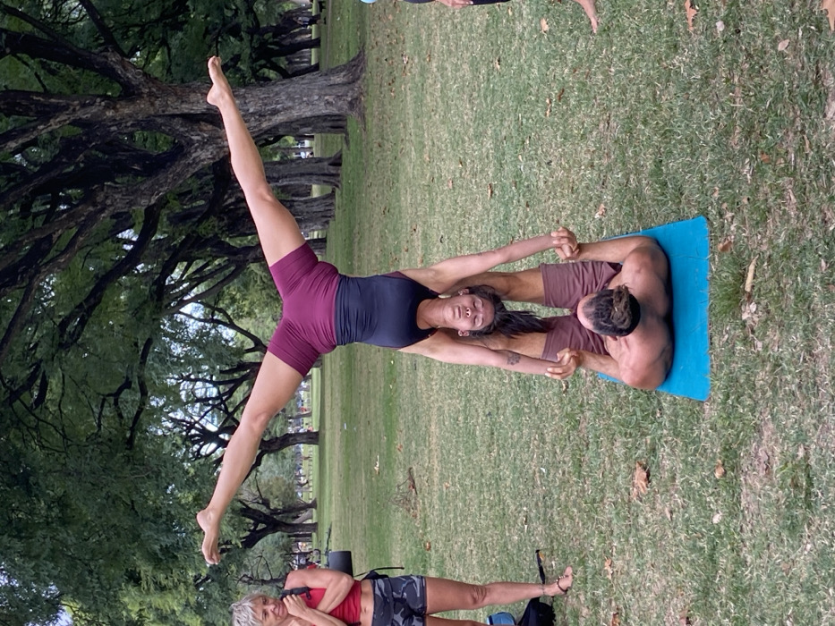 An outdoor scene depicts three individuals engaged in physical activity within a park setting, likely the Bosques de Palermo in Buenos Aires, Argentina.
In the foreground, a male and a female perform an acroyoga pose on a blue yoga mat placed on green grass with scattered brown leaves. The male, shirtless with dreadlocks and wearing dark red athletic shorts, lies supine on the mat. His legs are bent and raised, supporting the lumbar area of the female flyer. His hands grip the flyer's hands. The female flyer, with dark hair and wearing a two-toned dark red and black athletic bodysuit, is inverted above the male. Her legs are spread wide in a horizontal "V" shape, and her arms are extended downwards, grasping the male's hands. Her head is inverted, facing the ground.
To the left, a third female with short grey hair, wearing a red tank top, camouflage-patterned shorts, and dark sandals, stands observing the acroyoga practitioners. She carries a dark backpack over her left shoulder, and her hands are positioned near her mouth.
The background consists of numerous mature trees with dark, textured trunks and dense green foliage, suggesting an established urban park environment consistent with the Bosques de Palermo. The lighting is even, indicating an overcast sky or diffused daylight.