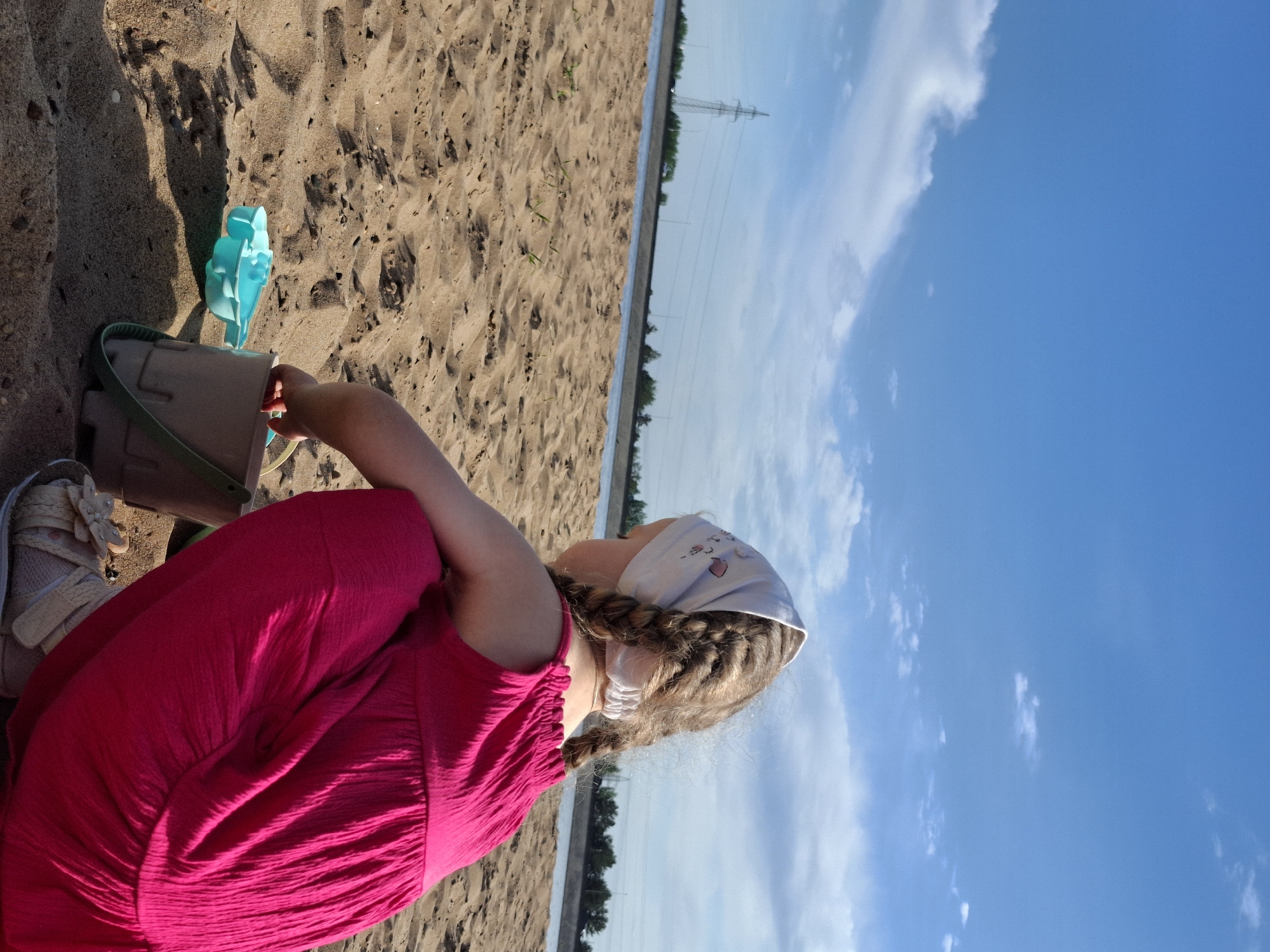 A little girl is playing on a sandy beach. She has blonde hair braided into pigtails and is wearing a white bandana. She's wearing a magenta dress, and her feet are clad in light-colored sandals. She is crouching near a sand bucket and a blue sand toy. The beach is a sandy expanse with some vegetation. In the background, across a body of water, are trees and utility poles. The sky is blue with scattered clouds. This is Noviy, Russia.