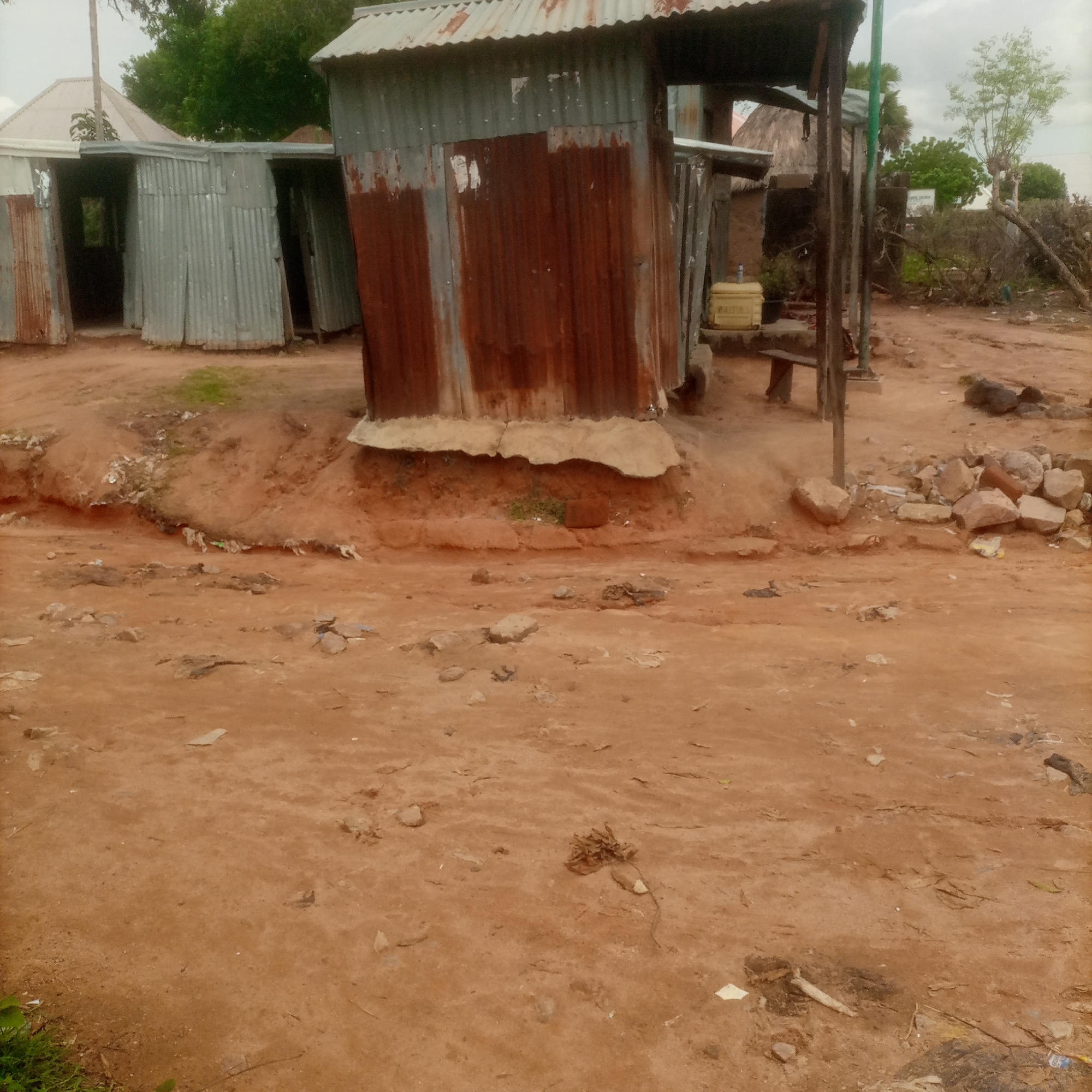 A weathered, corrugated metal shack stands in the foreground, its rusty panels contrasting with the lighter-colored tin roof. To the left of this shack, another similar structure is visible, appearing to be a smaller, attached room. The ground in front of the structures is a reddish-brown dirt path, scattered with stones and debris. In the background, a thatched roof structure is partially visible, suggesting a more traditional dwelling. Green foliage and trees can be seen behind the structures, and the sky is overcast. A yellow water container sits on a small platform next to the shack.