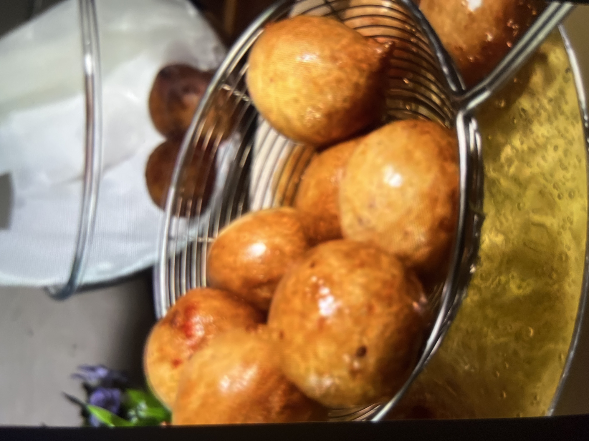 The image shows a close-up of golden-brown, fried dough balls, likely mandazi or puff-puff, being drained in a metal strainer over a pan of hot oil. The dough balls are round and have a slightly irregular, bumpy surface. Some have a reddish-brown hue, indicating they are well-fried. In the background, a glass bowl lined with white parchment paper contains more of the fried dough balls. A blurred object that appears to be a small arrangement of purple flowers and greenery is visible in the upper left corner. The overall lighting suggests an indoor setting, possibly a kitchen, with a focus on the cooking process. The scene does not provide any specific information that would allow for a determination of its location in Abuja, Nigeria.