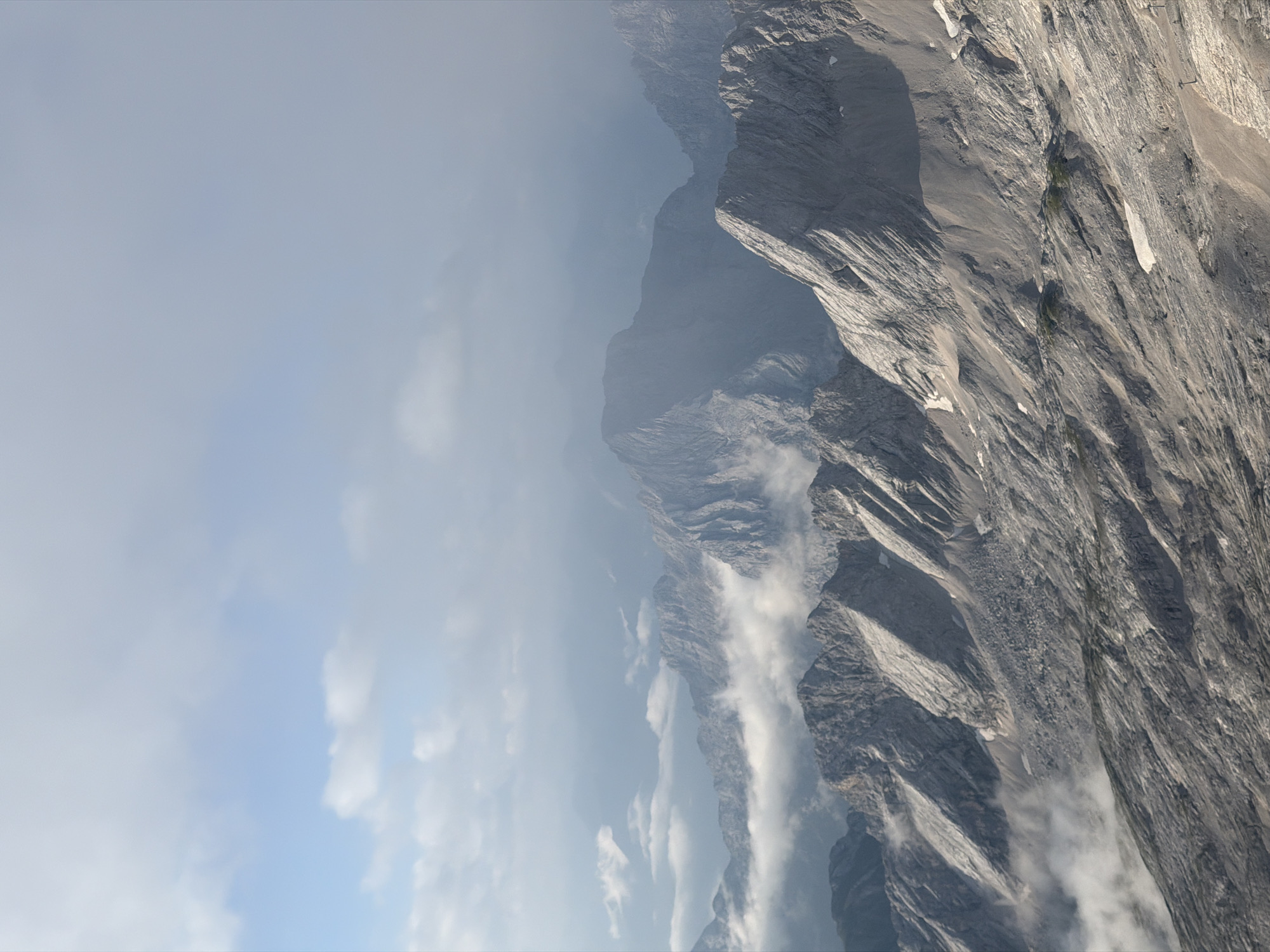 This is a vertical, eye-level, medium shot capturing a panoramic view of the Zugspitze mountain range near Grainau, Germany. The foreground is dominated by a rugged, rocky slope with visible scree and patches of snow. Jagged rock formations rise from the slope, with sharp ridges and textured surfaces. In the middle ground, several other mountain peaks emerge from a layer of wispy clouds. These mountains are also characterized by their rocky, eroded appearance, with steep inclines and visible geological layers. Further in the background, more distant mountain peaks are shrouded in haze and mist, blending into the sky. The sky above is a pale blue, with scattered white clouds, some of which are thin and wispy, while others are more substantial and appear to be drifting. The overall lighting suggests either early morning or late afternoon, with a soft, diffused light illuminating the scene. There are no people or specific objects like buildings or vehicles visible in this natural landscape. The environment is alpine, with a focus on the grandeur and raw beauty of the mountains.