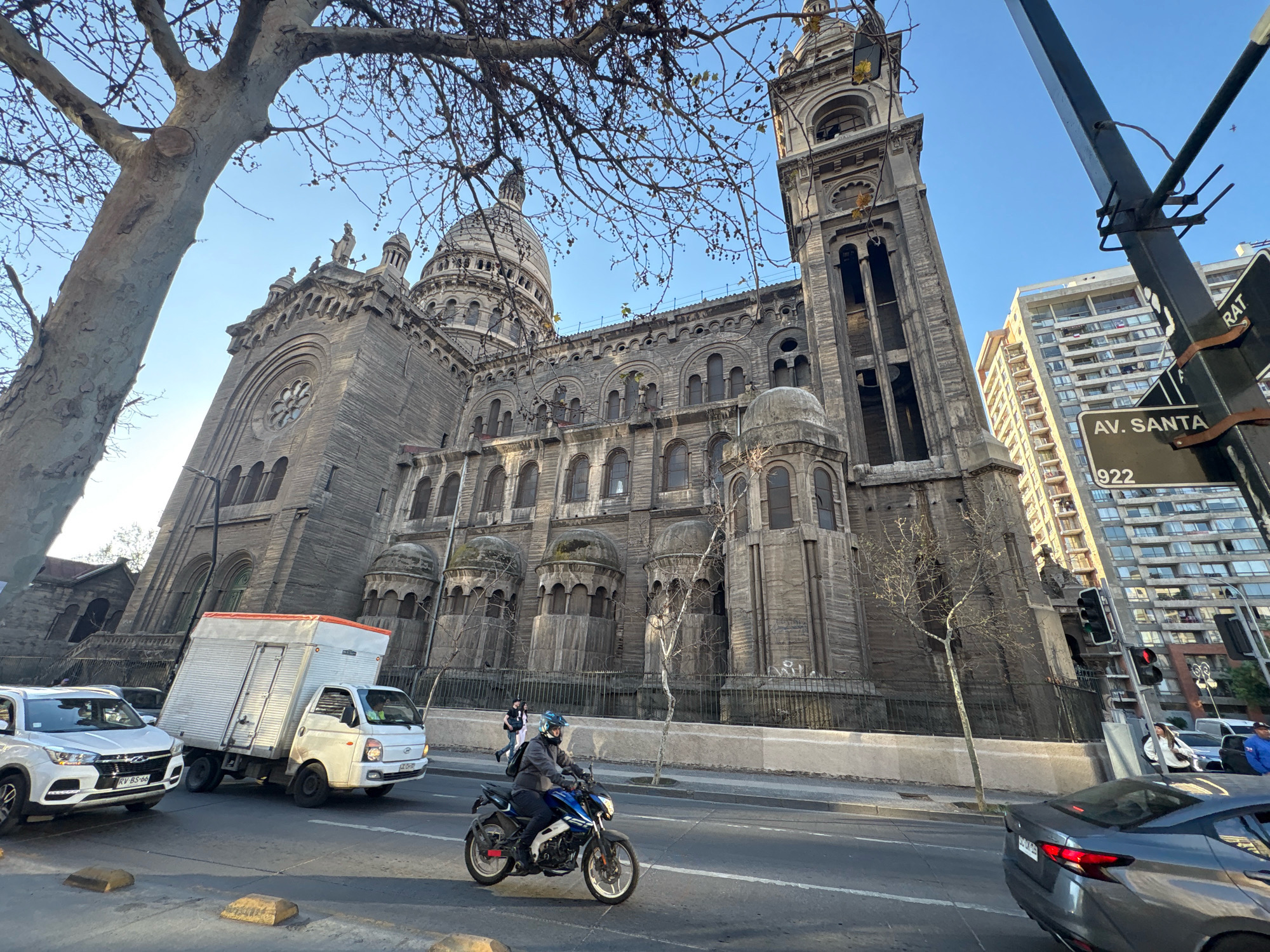 The image shows a view of the Metropolitan Cathedral of Santiago, Chile, from a street level perspective. The large, stone cathedral with its prominent dome and bell tower is the main subject, seen partially obscured by the branches of a large tree in the foreground. Several vehicles are on the street, including a white van, a white SUV, a grey car, and a motorcycle with a rider. People are visible walking and riding. Street signs for "AV. SANTA" and "922" are also visible. The scene appears to be on a clear day with a blue sky.