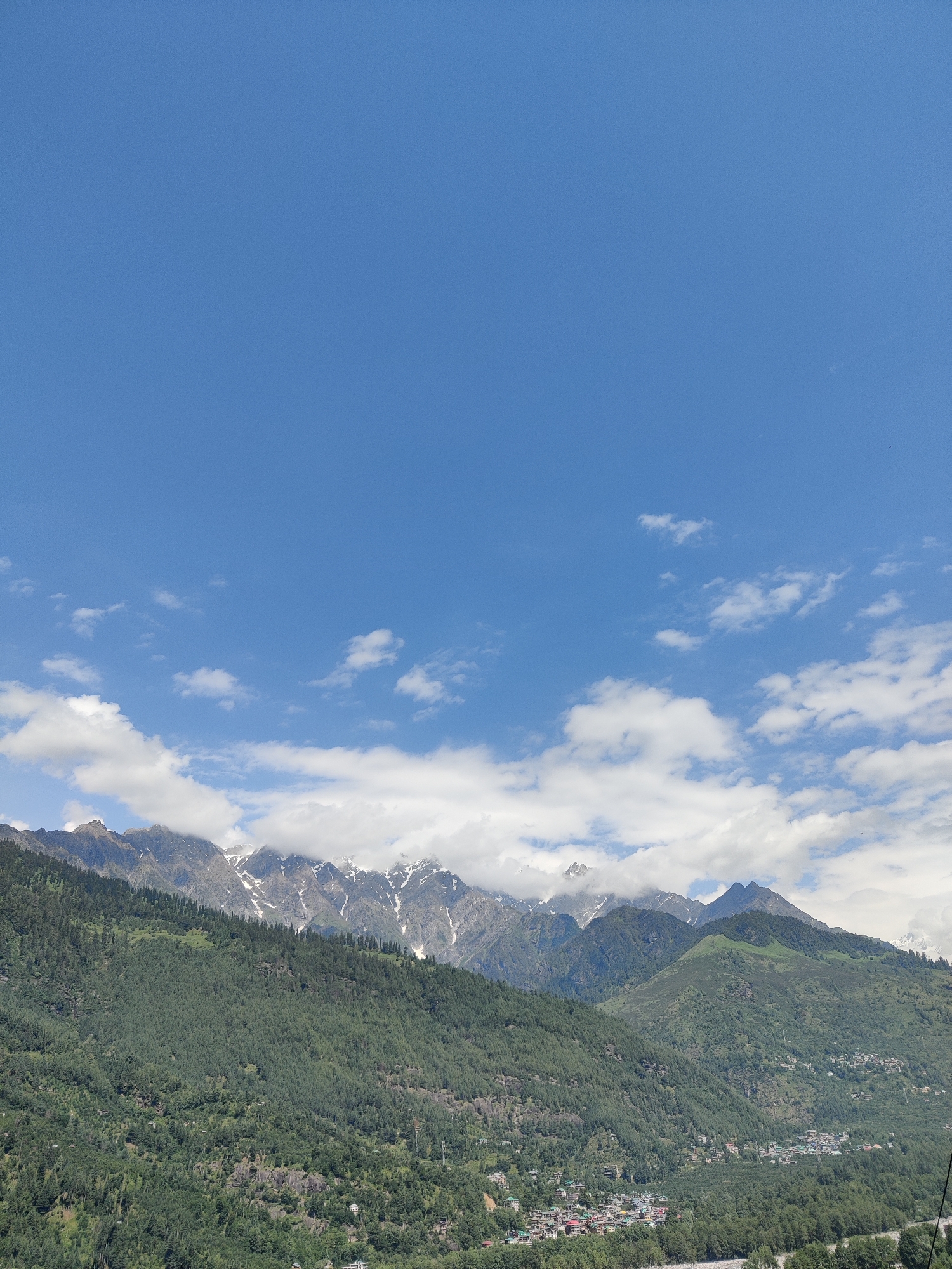 The image depicts a scenic view of a valley in Vashisht, India. The foreground is dominated by lush green mountains covered in dense vegetation, with scattered small houses and buildings visible on the slopes, suggesting a village or small town. The middle ground features more mountainous terrain, with some areas appearing rocky and others covered in trees. The background showcases a range of majestic snow-capped mountains, partially obscured by clouds, under a clear blue sky dotted with white, fluffy clouds. The overall scene conveys a sense of natural beauty and tranquility.