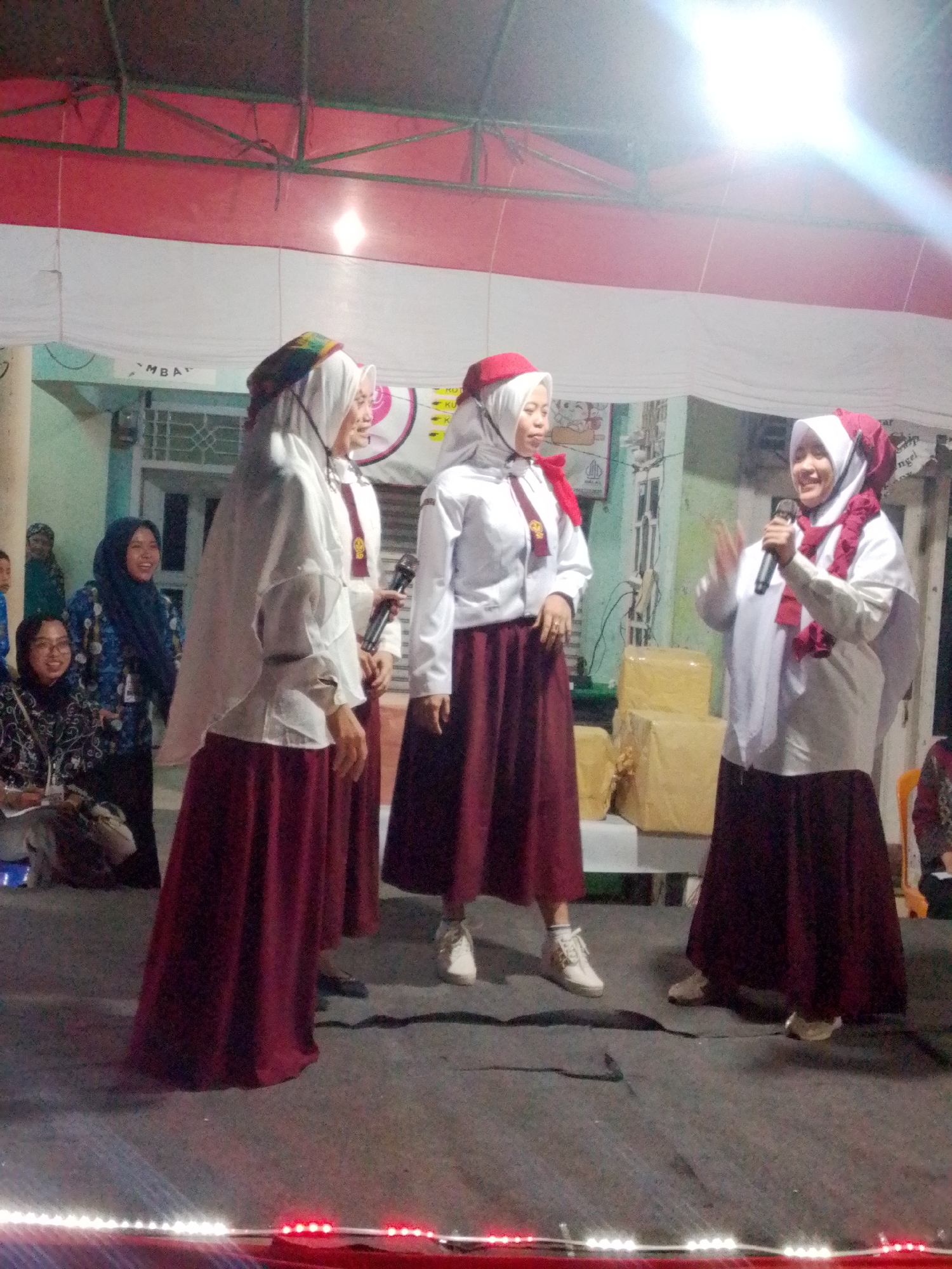 The image depicts an outdoor event, possibly a school performance or ceremony, taking place in Bandar Lampung, Indonesia. Four young women, all wearing maroon skirts and white long-sleeved tops with maroon hijabs, are the central figures. Three of them are standing on a dark carpeted area, with one holding a microphone and speaking, appearing to be leading the group or addressing an audience. The backdrop includes a white and red striped banner, likely representing the Indonesian flag, and some buildings. Several other individuals are visible in the background, including women sitting and watching the event. The scene is illuminated by overhead lights, suggesting it is evening or held under a covered area. In the foreground, there is a string of decorative lights.