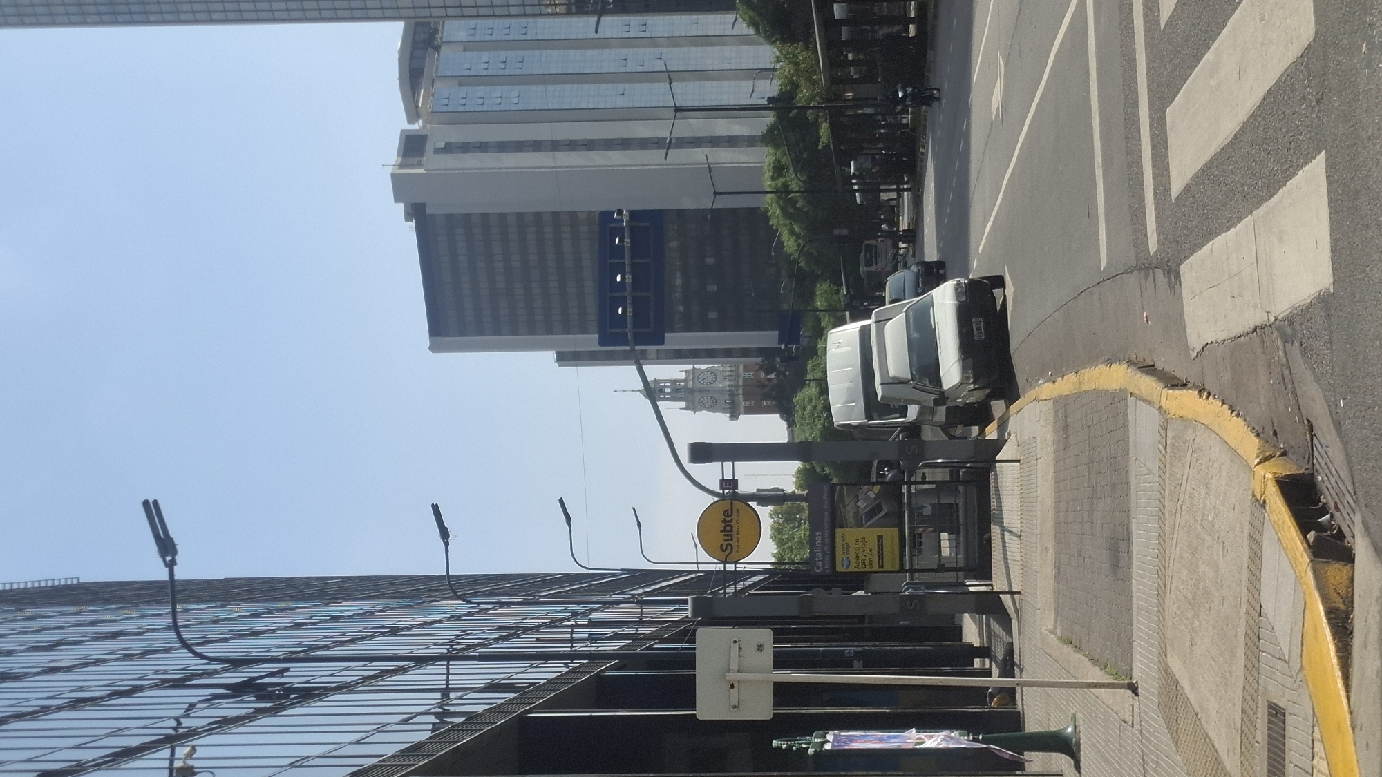 The image shows a street scene in Buenos Aires, Argentina. The photograph is taken at street level and is rotated. Prominent features include a "Subte" (subway) sign, a building with a glass facade, a crosswalk, several white vans, and a clock tower in the distance. The street is paved and has a yellow curb. There are streetlights lining the sidewalk. Several buildings are visible in the background, including modern high-rises. The sky is clear and blue. The location is likely near the Catalinas area, as indicated by signage in the image.