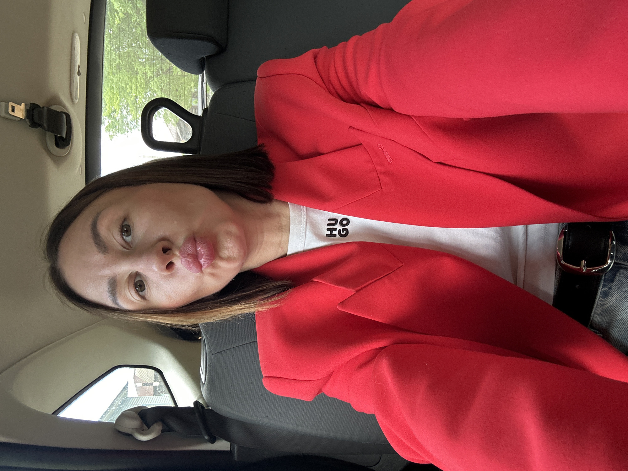 A woman with brown hair is seated in what appears to be the back of a vehicle, possibly a car, given the presence of a seatbelt and headrest. She is wearing a red blazer over a white t-shirt and a belt with a silver buckle. Her facial expression includes pursed lips. Outside the vehicle window, trees are visible, suggesting an outdoor environment. Without further details, it's impossible to verify if the location is Votkinsk, Russia.