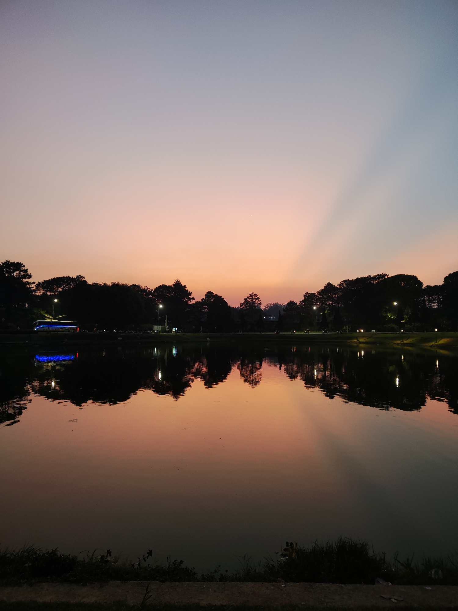 A photograph depicts a serene lake at dusk in Da Lat, Vietnam. The sky is a gradient of peach and light blue, with crepuscular rays visible in the upper right. The lake reflects the colors of the sky and the silhouetted trees lining the shore. Several lampposts are visible along the perimeter. A blue bus stands illuminated on the left side of the lake. The foreground shows the bank of the lake with grass and a concrete edge. The overall scene is one of tranquility and natural beauty.