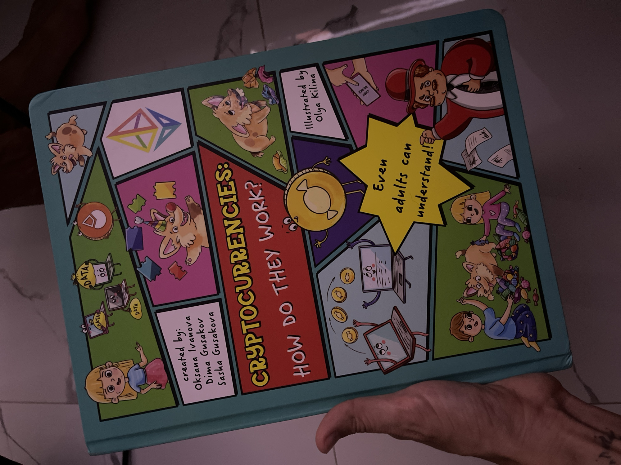 The image shows the cover of a children's book titled "CRYPTOCURRENCIES: HOW DO THEY WORK?". The cover is vibrantly illustrated with cartoonish characters and graphics related to technology and finance. In the top left corner, a girl is shown with laptops and the word "DATA". To her right, there are animated characters resembling dogs and a circular orange object with an eye. In the center of the upper half, an Ethereum logo is displayed within a geometric shape. The lower half of the cover features animated laptops with coins floating between them, a character resembling a piece of candy with arms and legs, and a section with text that reads "Even adults can understand!". To the right of this, there is text indicating "Illustrated by Olya Kilina". Below this, a boy and a dog are depicted surrounded by candy. Further to the right, a man in a suit and hat is shown with papers. The book is held by a person's hand on the left side of the image, obscuring a small portion of the cover. The background is indistinct, suggesting an indoor setting. The presence of the book and the hand holding it, along with the faint shadows, do not provide specific geographical markers to indicate a location in ตำบลเกาะพะงัน, Thailand.