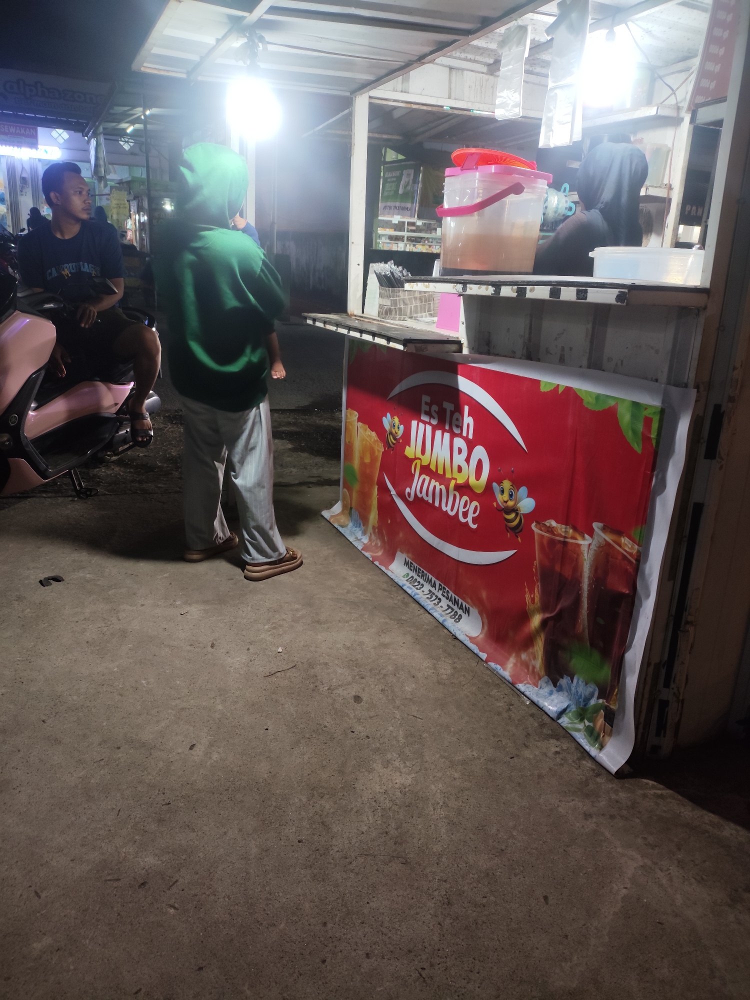 The image depicts a street-side food stall in Palembang, Indonesia, at night. A man is seated on a motorcycle, looking towards the left. Another person, wearing a green hoodie and grey striped pants, stands facing a counter. Behind the counter, a large dispenser filled with a brown liquid and a red lid is visible. A banner is displayed on the front of the stall, featuring the text "Es Teh Jumbo Jambee" with bee illustrations and images of iced tea. A phone number, "0823-7573-7788", is also printed on the banner, suggesting it is a contact for orders. The environment appears to be a dimly lit outdoor area with a concrete ground.