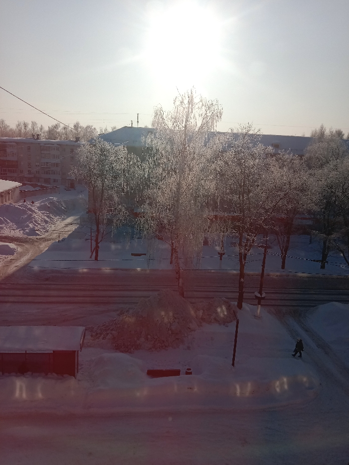 The image captures a bright, sunny winter scene in what appears to be an urban or semi-urban environment, viewed from an elevated perspective, possibly from a window. The sky is very bright, almost overexposed, with the sun radiating prominently in the upper center, creating a strong lens flare or reddish tint across the lower portion of the image.
The ground is heavily covered in white snow, which also blankets rooftops and forms significant piles along roadsides, suggesting recent snowfall and snow plowing. Numerous bare trees, likely deciduous, are scattered throughout the scene. Their branches are beautifully coated with a thick layer of frost or ice (hoarfrost/rime ice), making them appear white and glistening under the sunlight. A tall, slender tree, possibly a birch, stands prominently in the mid-ground, its delicate, frost-laden branches catching the light.
In the background, multi-story residential buildings with snow-covered roofs are visible. Some utility poles and power lines crisscross the upper part of the frame. A main road runs horizontally across the middle ground, appearing mostly clear but with snow piled along its edges. Smaller paths and driveways, also snow-covered, branch off from it.
On the right side of the image, towards the lower quarter, a lone individual is visible walking on a snow-covered path. They are dressed in dark winter clothing. In the lower-left corner, a small, single-story building, possibly a garage or shed, with a snow-covered roof, is partially visible.
The overall impression is one of a crisp, cold, and beautiful winter day with abundant snow and bright sunshine. The reddish light flare in the foreground adds a unique photographic artifact to the scene. No discernible text is visible in the image.