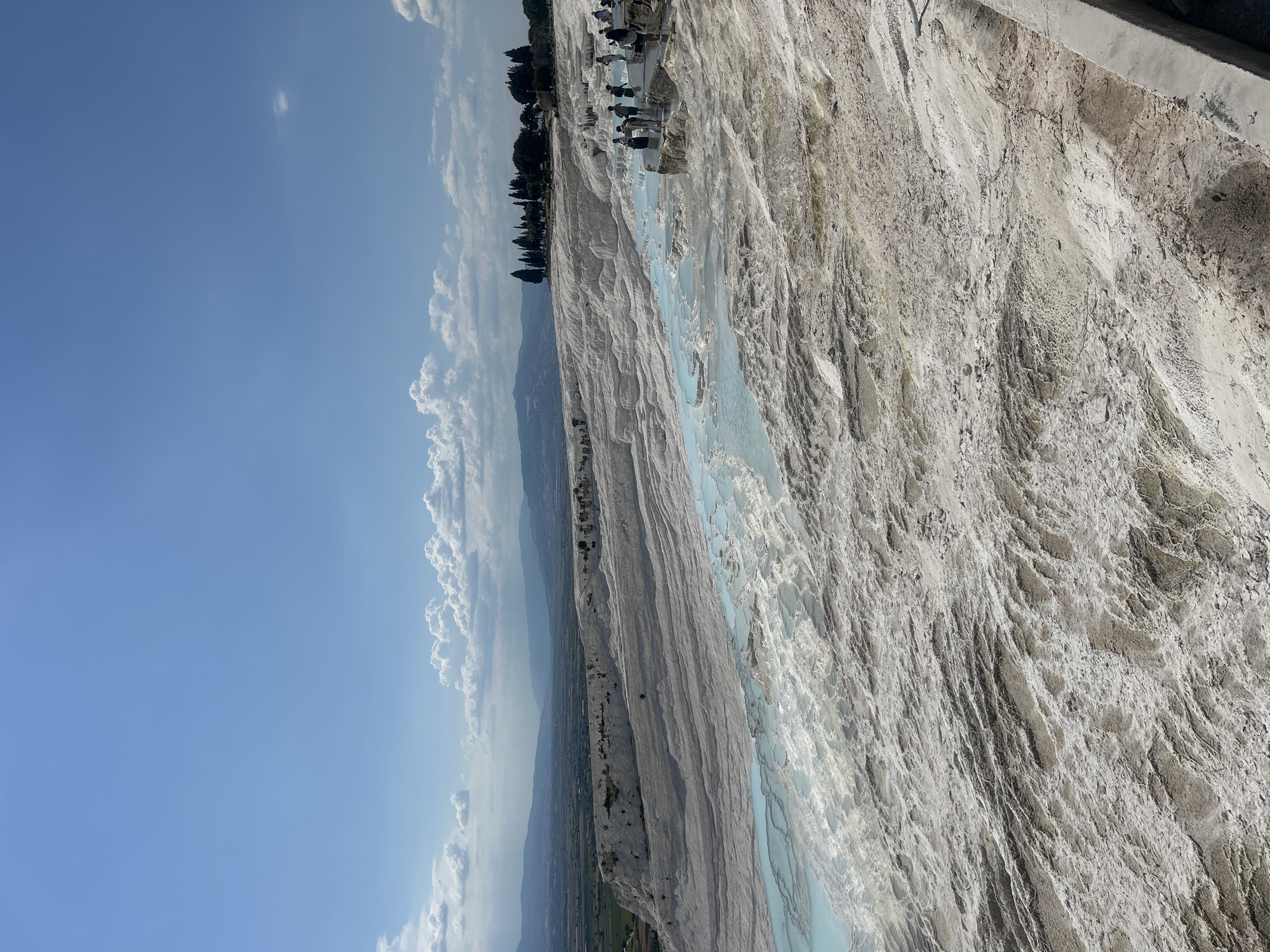 The image depicts Pamukkale, Turkey, a natural site with travertine terraces and hot springs. The terraces are white, formed by calcium carbonate deposits from the mineral-rich waters. Pools of turquoise water fill the terraces. At the top of the terraces, there are trees and people. The sky is blue with white clouds. In the distance, there is a landscape with mountains and a plain.
