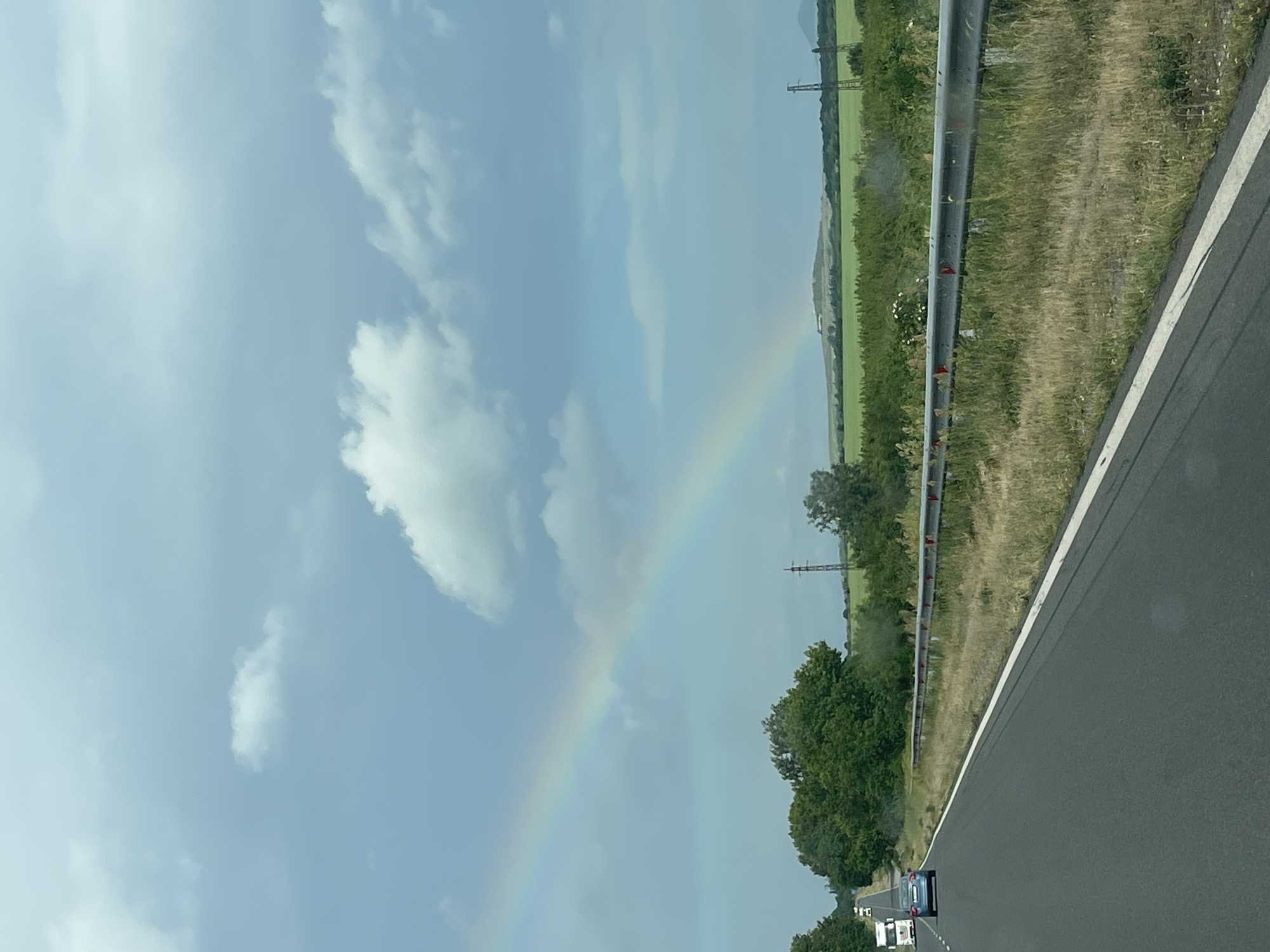 The image shows a view from a highway near Seliminovo, Bulgaria. A rainbow arcs across the sky on the left side of the frame. To the right of the rainbow are fields and some scattered trees. A metal guardrail runs alongside the highway, and a white stripe marks the edge of the road. In the bottom right corner, the asphalt road stretches into the distance. Two vehicles are visible on the road, one appears to be a white truck and the other a blue car. Power lines can be seen crossing the fields. The sky is mostly cloudy.