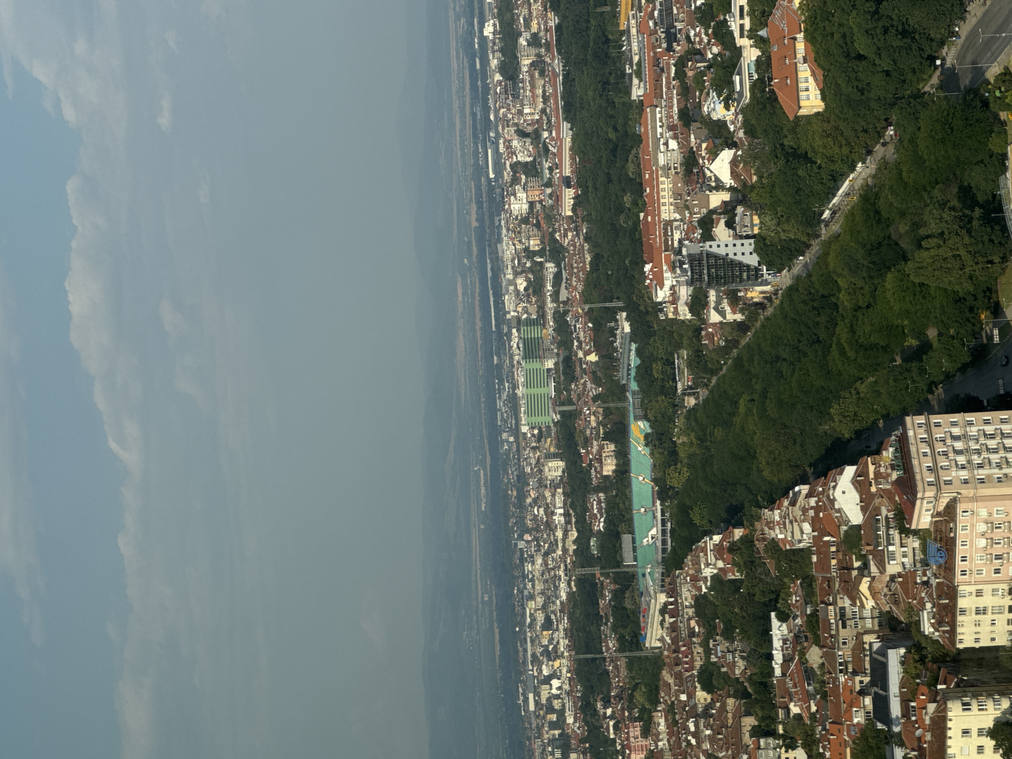 This is a high-angle aerial view of a cityscape in Sofia, Bulgaria. In the foreground, a dense urban area with residential buildings featuring red-tiled roofs dominates the scene, interspersed with green trees and a road running diagonally. Further into the midground, a large green stadium with seating is visible, surrounded by more trees. Beyond the stadium, the city extends outwards with a mix of buildings of varying heights and colors. In the far background, a hazy landscape of rolling hills or mountains stretches across the horizon under a sky with scattered clouds. The overall impression is a sprawling urban environment with significant green spaces.