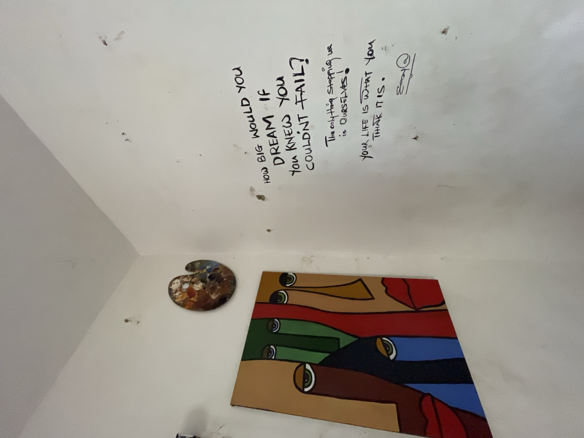 The image depicts an interior corner of a room, likely an artist's studio, located in Ado, Nigeria. The walls are painted a light, neutral color and show signs of wear and tear, with small marks and imperfections visible.
Dominating the left side of the frame is a colorful abstract painting of faces. The painting features bold geometric shapes and vibrant colors such as red, green, blue, and brown, with prominent eyes and lips rendered in a stylized manner. Above and slightly to the right of the painting, a wooden artist's palette, splattered with various paint colors, is affixed to the wall.
The right side of the wall is adorned with black, handwritten text. The largest inscription, in a bold, angular script, asks: "HOW BIG WOULD YOU DREAM IF YOU KNEW YOU COULDN'T FAIL?". Below this, in a smaller, more cursive font, it reads: "The only thing stopping us is OURSELVES!". Further down, another message states: "YOUR LIFE IS WHAT YOU THINK IT IS." A stylized signature, appearing as "Easy @", is located at the bottom right of the inscribed text. Scattered on the wall are a few dark spots, possibly dirt or paint splatters. The ceiling is visible at the top of the image, also appearing to be a light color.