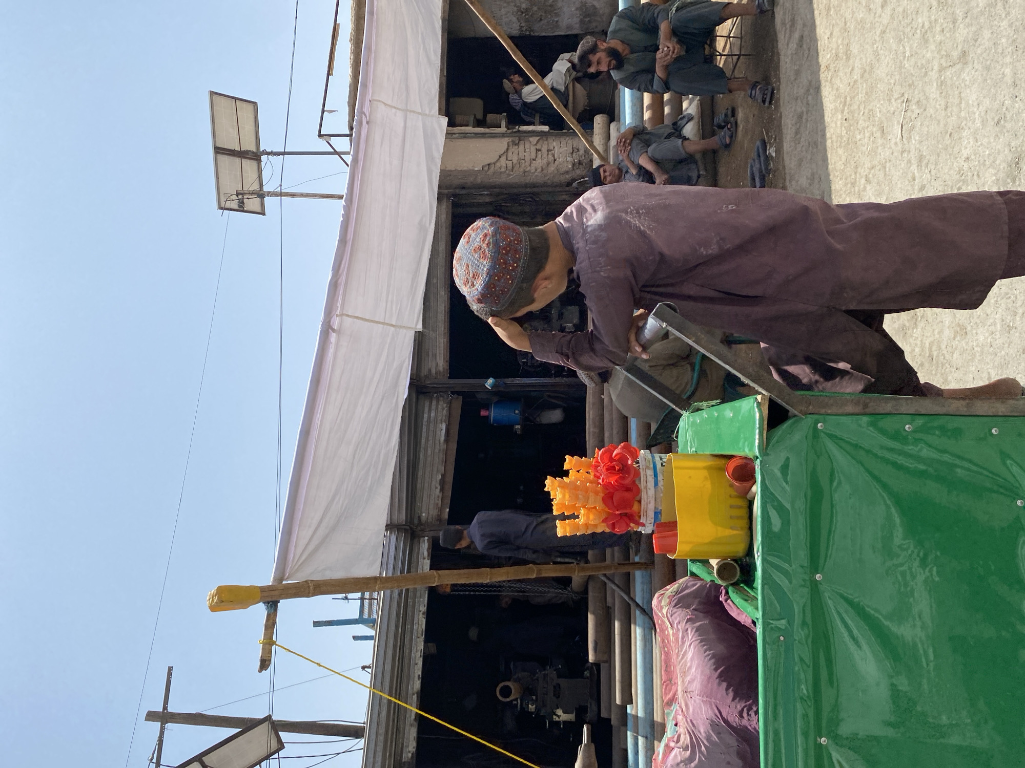 In the city of Kandahar, Afghanistan, a young boy wearing a maroon tunic and a patterned cap stands on the left, facing away from the camera, his hand on his head. In front of him is a green cart covered with a green tarp. On the cart, a yellow bucket holds red and orange objects resembling flowers or ice cream cones. A purple cloth lies on the left side of the cart. Behind the cart, a shop with a corrugated metal facade has its shutters open, revealing a dark interior where two men are visible. To the right of the shop, three men are seated on a bench or low structure. One man is in the foreground on the right, wearing a dark green tunic and pants, with a beard and short dark hair. He is looking to the left. Another man, wearing a dark tunic, is seated behind him. A third man, in a light-colored tunic, is seated further back and to the left. The ground is dusty and uneven. Overhead, a white tarp is stretched across poles, providing shade. A solar panel is visible on a pole in the background.