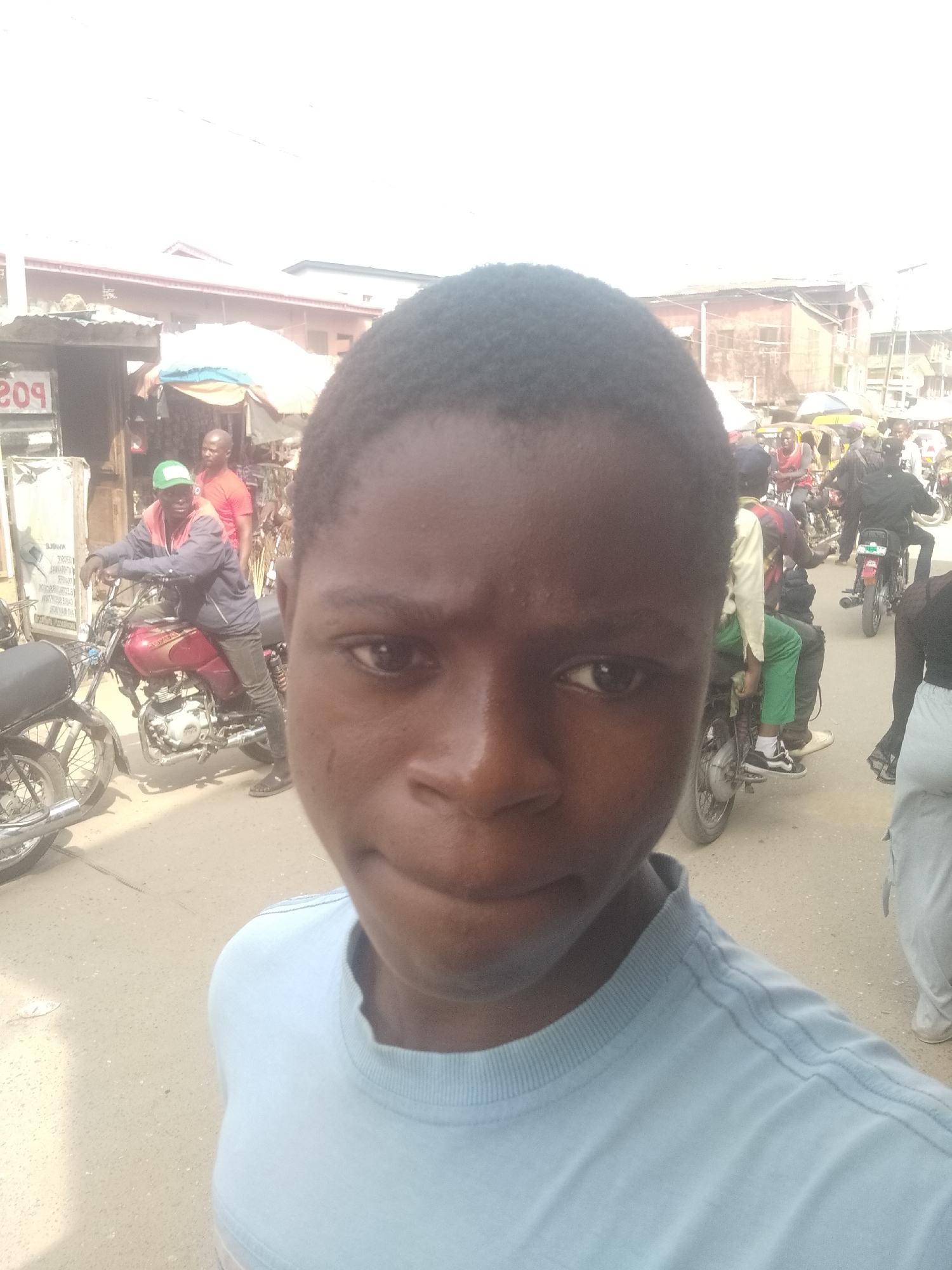 The image shows a medium close-up of a young, dark-skinned male wearing a light blue t-shirt. The background reveals a crowded street scene in an urban location. There are numerous motorcycles and people visible in the background. There's a small stall with a sign with the number "209" partially visible. The scene appears to be outdoors in daylight. The location is Unknown, Unknown.