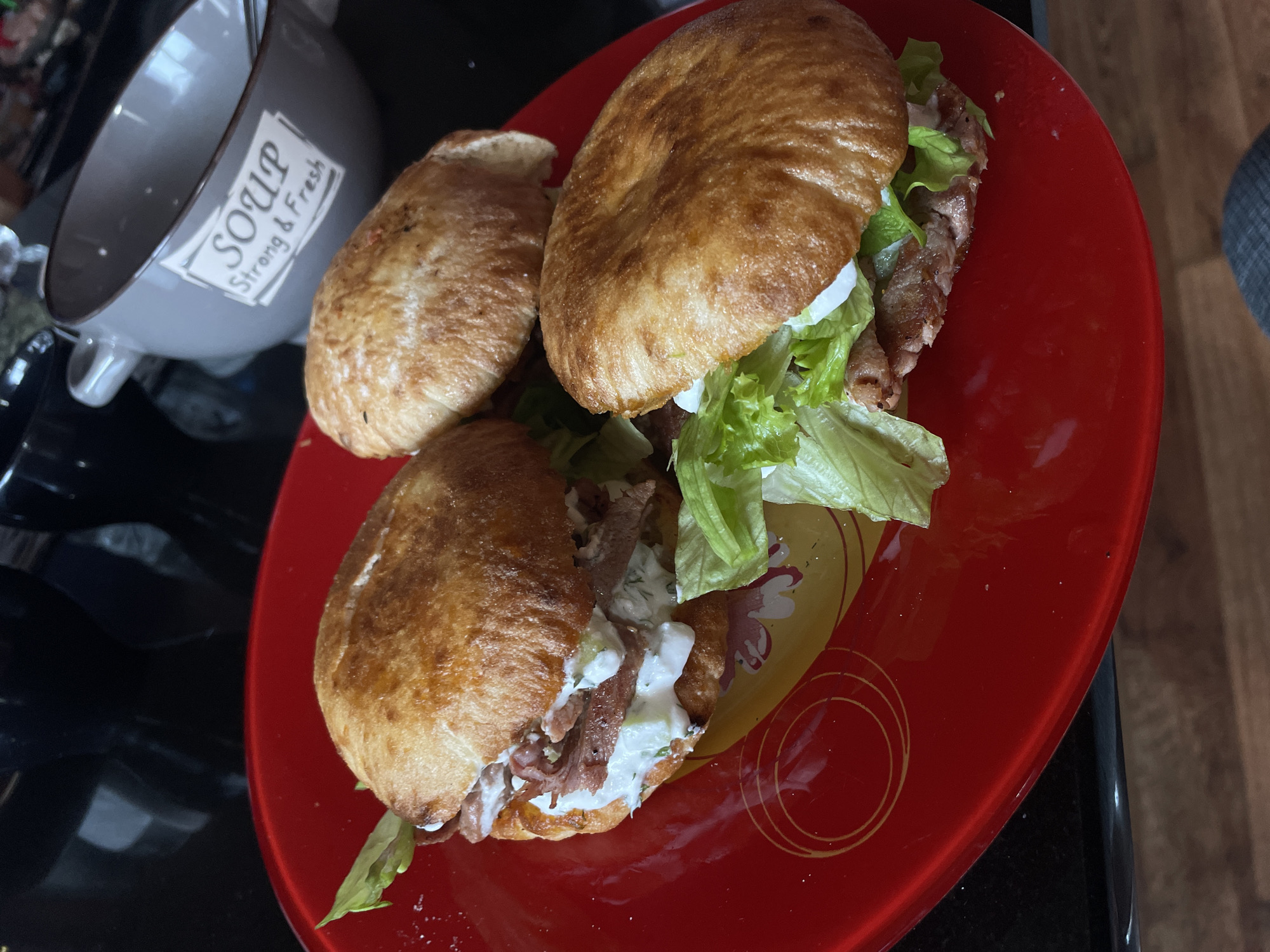 The image depicts a close-up shot of a red plate containing what appear to be two and a half sandwiches, likely gyros or similar pita-based sandwiches. The sandwiches are composed of toasted pita bread, lettuce, meat (possibly lamb or beef), and a white sauce (potentially tzatziki). The plate has a decorative pattern with concentric circles. In the background, a gray soup bowl with the text "SOUP Strong & Fresh" is visible. The items are situated on a black surface, possibly a countertop. The photo appears to be taken indoors, with wooden flooring partially visible in the corner. No information is available to confirm location.