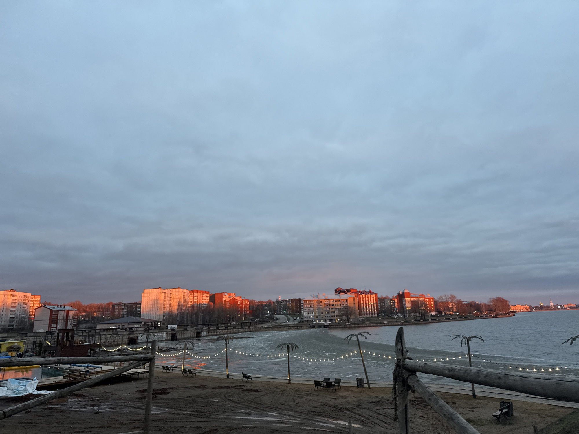 This wide-angle photograph captures a waterfront scene in Votkinsk, Russia, during the golden hour of either sunrise or sunset. The sky above is largely overcast with grey clouds, creating a muted backdrop, but a warm, orange light from the low sun dramatically illuminates the upper portions of multi-story buildings lining the distant shore.
In the foreground, a sandy or muddy beach area occupies the lower third of the frame. A rustic wooden fence constructed from logs runs diagonally across the right side. Several stylized, dark "palm tree" cutouts are planted in the sand, and string lights are gracefully suspended between them, forming illuminated garlands along the water's edge. Dark outdoor tables and chairs are scattered on the sand, suggesting a recreational or dining area, though no people are visible. On the far left, some colorful objects, possibly small boats or water recreation equipment, are partially covered with blue tarps.
The body of water in the middle ground shows signs of cold temperatures, with noticeable ice floes or frozen sections near the shore, particularly in the central part of the image. A bridge-like structure is visible stretching across the water, connecting to the opposite bank. The cityscape beyond consists of numerous residential and possibly commercial buildings, their windows and facades reflecting the warm, red-orange glow. Behind these structures, sparse, bare trees indicate a cold season.
The overall mood is quiet and serene, characterized by the striking contrast between the cool, muted tones of the grey sky and partially frozen water, and the vibrant, warm light hitting the urban structures. There is no visible text in the image, and no specific human activities are taking place; the prepared leisure area appears empty at this time.