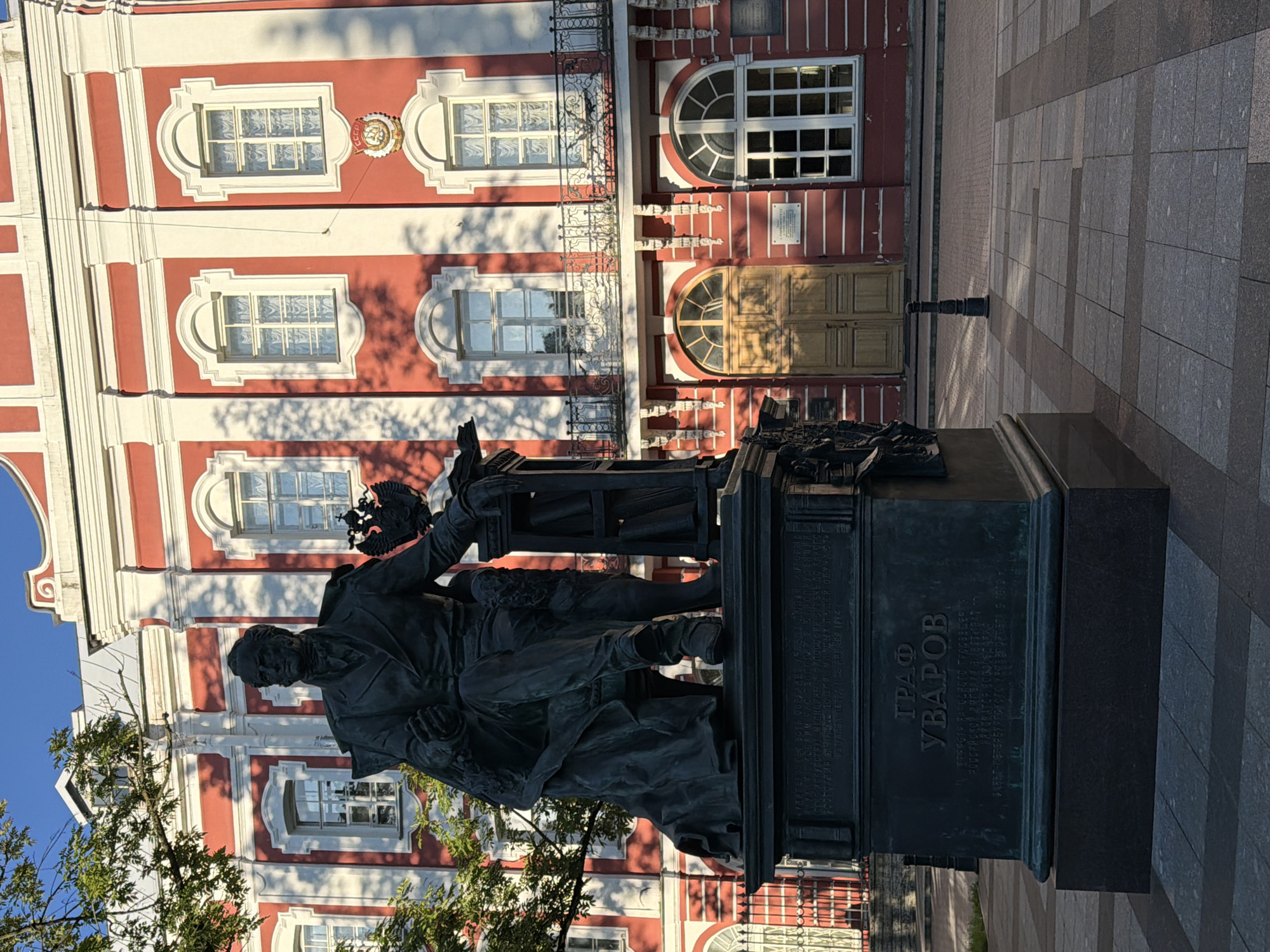 The image shows a bronze statue of a seated man in a suit in front of a red and white building in Saint Petersburg, Russia. The building appears to be a historic structure with arched windows and decorative architectural elements. The statue is mounted on a large, dark pedestal with inscriptions. A tree with green leaves is partially visible to the left of the statue. The ground in front of the building is paved with large square tiles. The sky is clear and blue. The lighting suggests it is either early morning or late afternoon. The statue is likely a monument to a prominent figure, with the inscription "ГРАФ УВАРОВ" (Count Uvarov) visible on the pedestal. This monument is located on the grounds of Saint Petersburg State University, in front of the Twelve Collegia building.