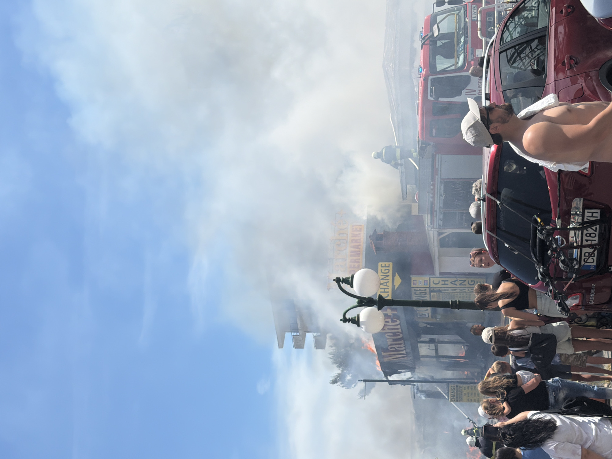The image captures a chaotic scene of a building fire in Nesebar, Bulgaria. Thick smoke billows into a bright blue sky, indicating it's daytime. Several red fire trucks are present, suggesting an active firefighting operation. A group of people are gathered, observing the fire, with some appearing to be pointing or looking concerned.
One of the most prominent figures is a shirtless man wearing a white baseball cap and sunglasses, with a white towel draped over his shoulders. He is looking upwards, seemingly towards the smoke. Other individuals, including women and children, are also visible in the foreground and background, some dressed casually in summer clothing.
Amidst the smoke and flames, there are signs that provide clues to the location. One sign reads "MARCHE" and another "SUPERMARKET," suggesting the fire is at or near a commercial establishment. There's also a yellow sign with an arrow and the word "CHANGE," likely indicating a currency exchange. A license plate on a red car visible in the image reads "CB 1824 TH".
The scene conveys a sense of urgency and alarm, with the dramatic presence of smoke and fire trucks. The onlookers' expressions, though not clearly discernible due to the image quality and distance, suggest a mix of concern and curiosity. The overall atmosphere is one of an unfolding emergency.