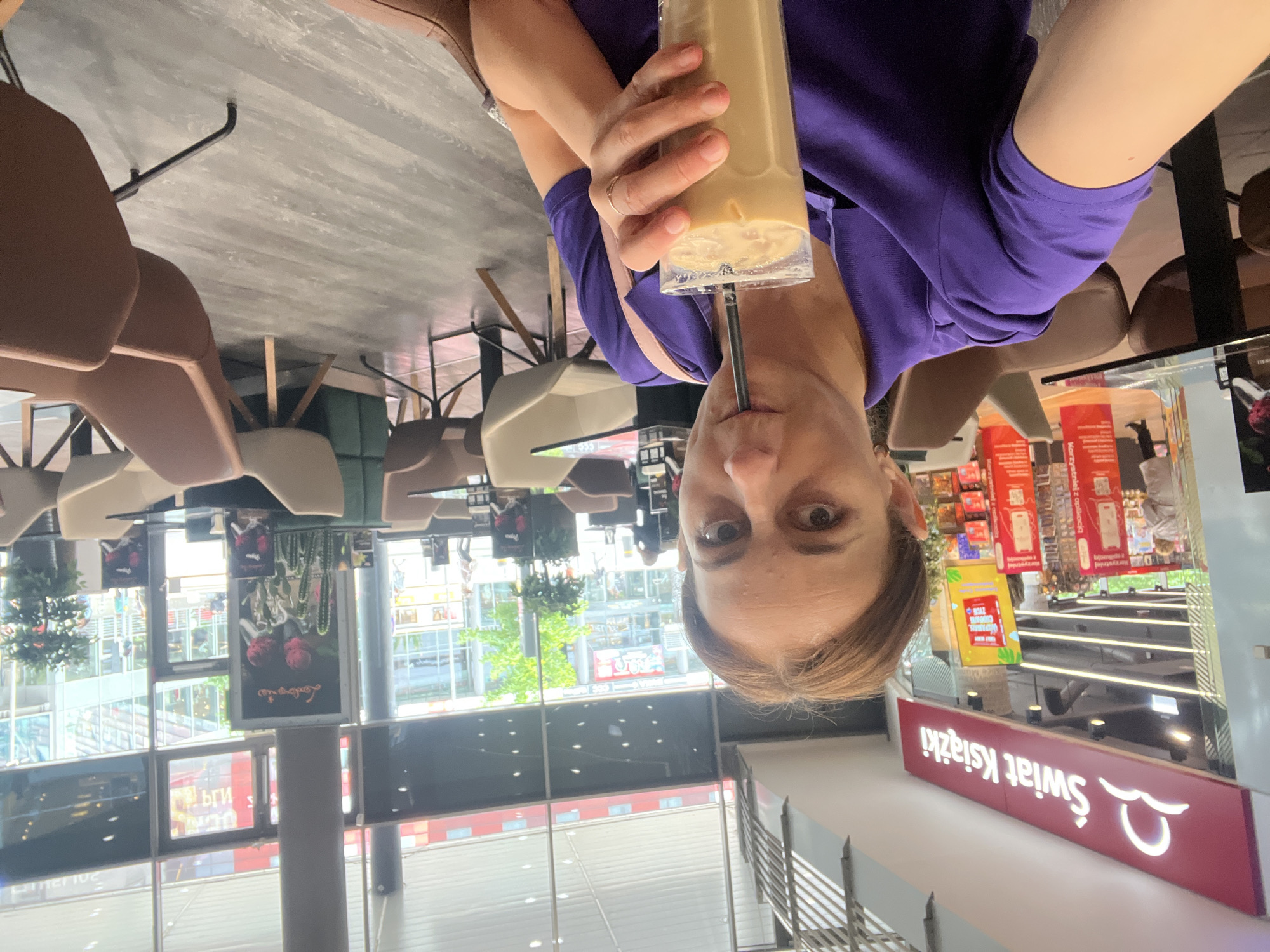 A woman is in the foreground of the image, holding a tall glass of iced coffee with a straw and drinking it. She is wearing a purple polo shirt. The background shows a modern interior with tables and chairs, suggesting a cafe or seating area within a larger establishment. To the left, a storefront with a sign reading "Świat Książki" is visible, indicating a bookstore. Large windows in the background reveal an urban environment with buildings and people outside.