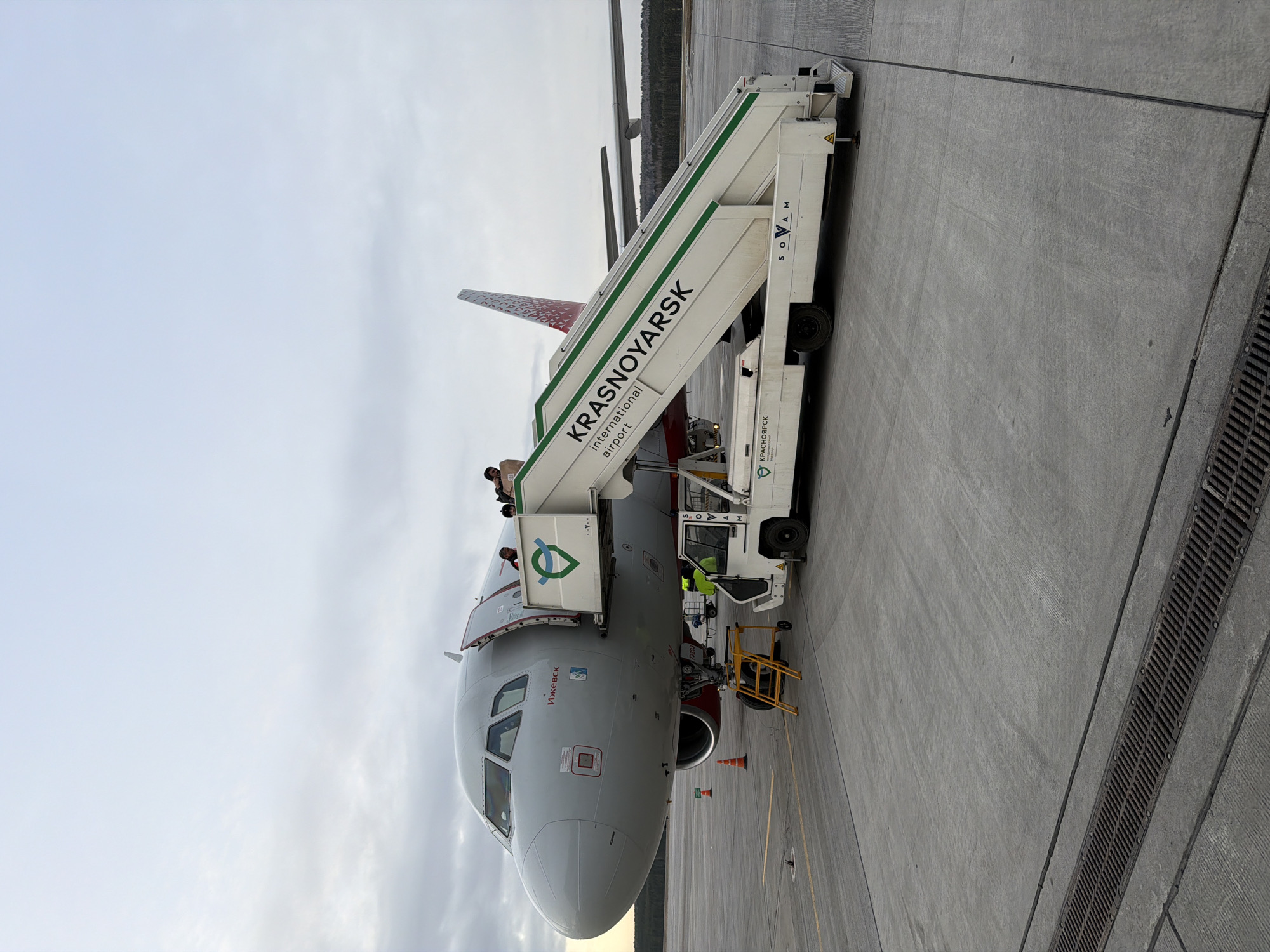 An airport scene in Емельяново, Russia, features an aircraft with a boarding stair vehicle connected to its front entrance. The plane's nose is prominently visible, exhibiting a gray-white color scheme with red accents near the engine area and tail. “Krasnoyarsk International Airport” is labeled on the boarding stairs. People are visible on the aircraft entrance with the boarding stairs. A smaller yellow utility vehicle is parked beside the stairs. The tarmac is gray concrete, and the sky is overcast.