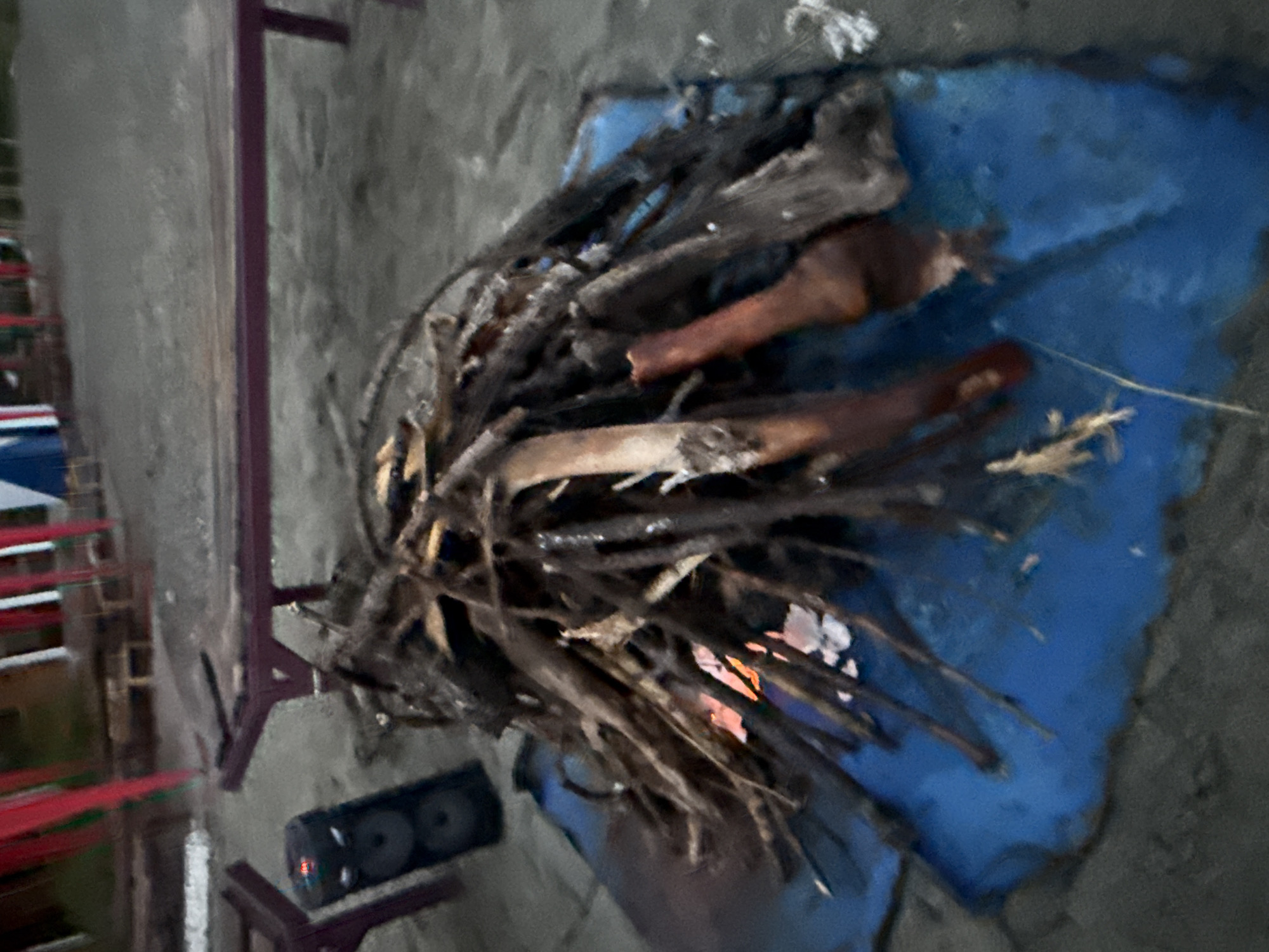 The image shows a bonfire on a blue tarp on a sandy surface, likely a beach or campground. The bonfire consists of a pile of dry branches and logs, with some embers glowing orange and white at the base. To the left of the bonfire is a black portable speaker. In the background, there are wooden benches and tables, suggesting an outdoor seating area. Red and green flags or banners are visible, possibly indicating a festive or celebratory setting. The overall environment appears to be outdoors, with a slightly overcast sky, typical of early evening or dusk. The location is identified as Sevan, Armenia, implying a setting near Lake Sevan, known for its beaches and recreational areas.