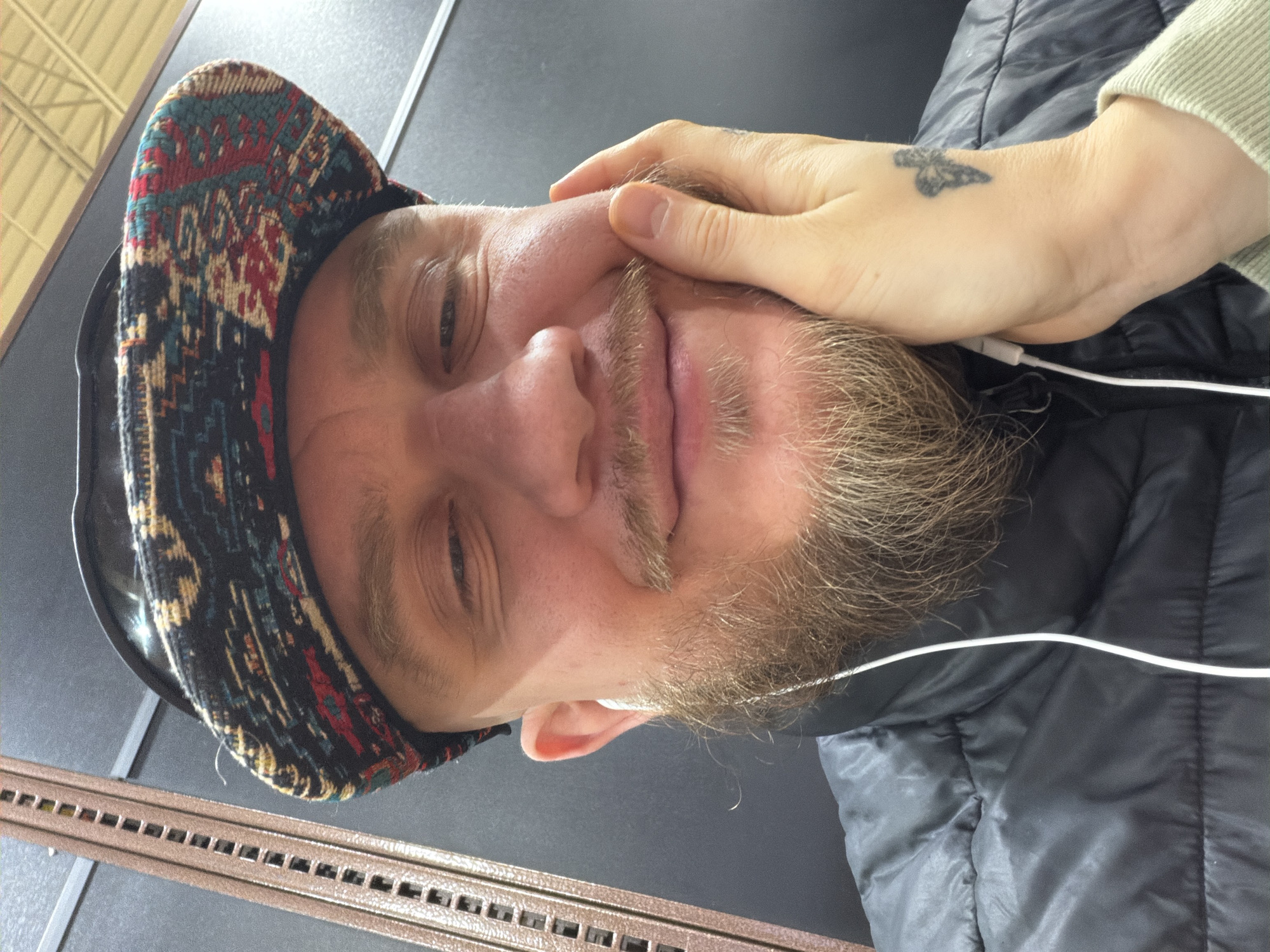 A close-up, slightly rotated headshot depicts a person with light skin, a beard, and wearing a patterned hat. Part of a hand with a butterfly tattoo is visible on the right side of the frame, touching the person's face. The person appears to be wearing a black puffer jacket and has white earbud wires visible. The background consists of dark gray panels and a brown, perforated metal structure along the bottom edge. Based on the visual details, it is inferred that the location is in Niles, United States of America.