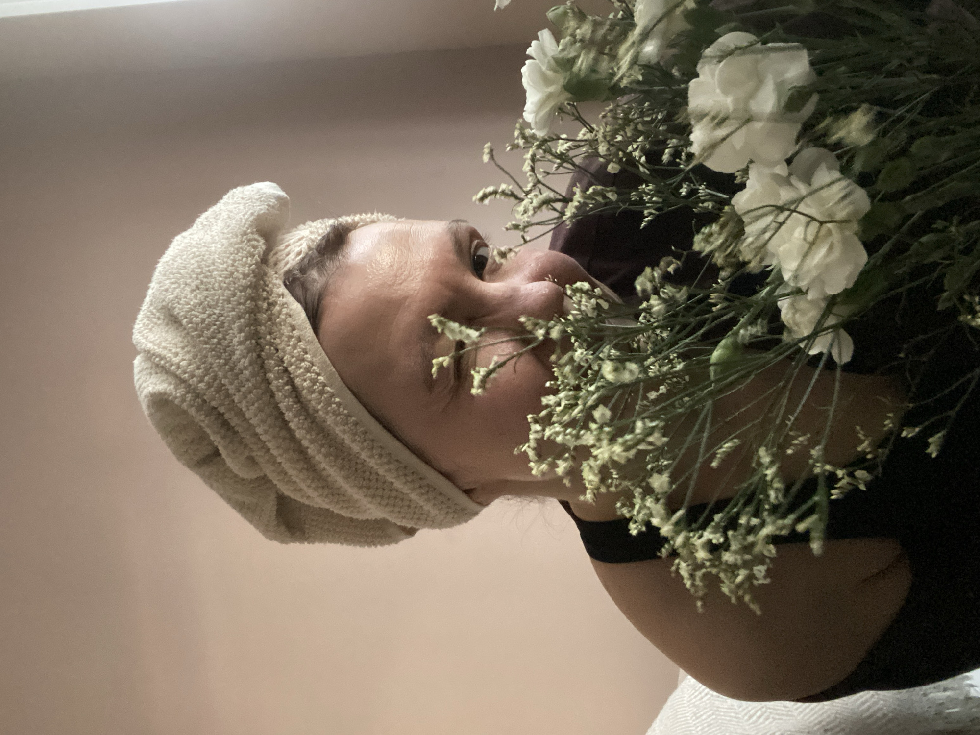 The image shows a close-up, rotated view of a person with a bouquet of flowers in an indoor setting. The image is oriented such that the person's head, wrapped in a light beige, textured towel, appears on the left, looking upwards and to the right.
The central subject is the person's face in profile, with their nose and mouth partially obscured by the dense foliage of the flower bouquet. Their right eye is wide open, gazing intently at the flowers. The skin appears light-toned. The towel wrapped around their head is a thick, cream or light beige color with a visible texture, possibly terry cloth or a similar material, suggesting recently washed hair.
The bouquet consists primarily of white flowers, identifiable as carnations, mixed with smaller, delicate clusters of what appears to be baby's breath or limonium, providing a contrasting green and white composition. The person is holding the bouquet close, seemingly smelling or closely examining the blossoms.
The background is a plain, light pink or beige wall, indicating an indoor environment. There are no other objects or details visible in the background to provide further context about the specific location within Chaikovsky, Russia. The lighting is soft and even, suggesting ambient indoor illumination. No text is visible in the image.