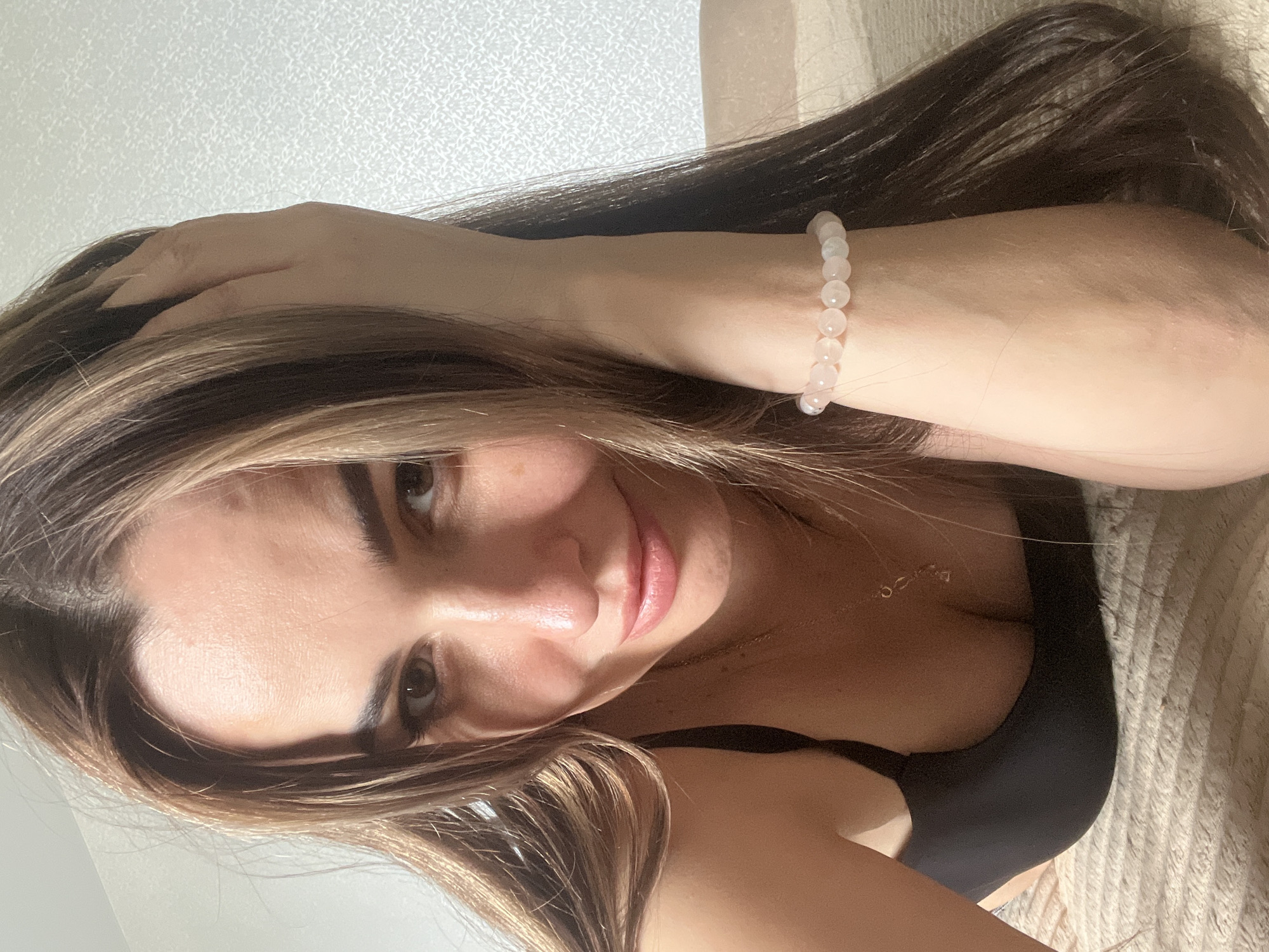 A selfie of a woman with brown hair, some highlights, and light skin. She is wearing a black top and a beaded bracelet on her left wrist. She is indoors. The city and exact location are unknown.