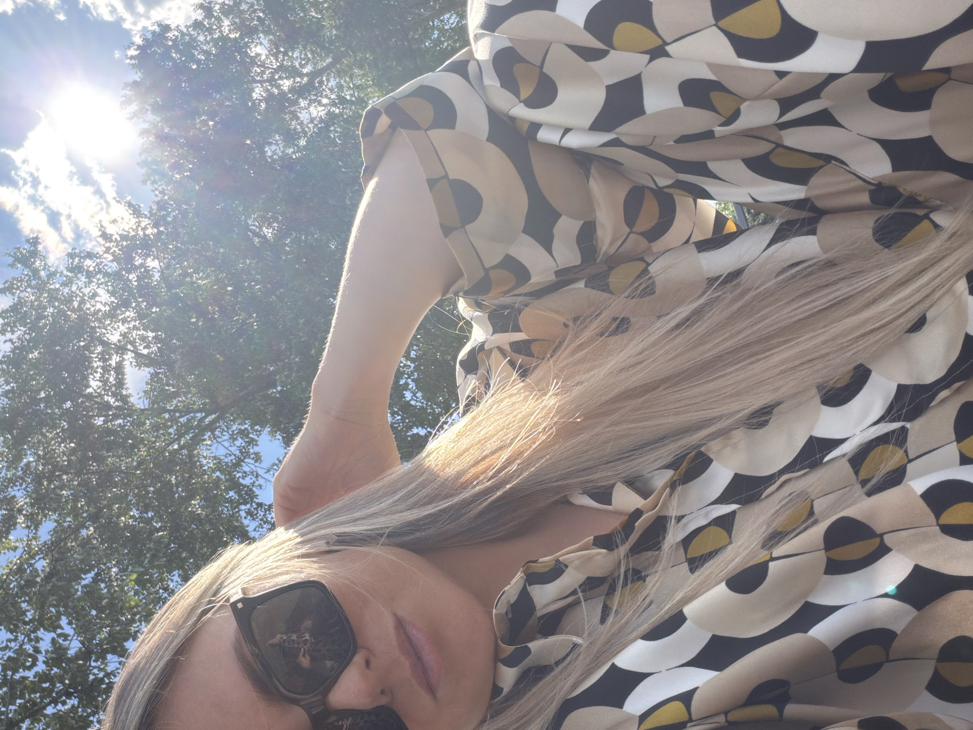 A close-up, slightly low-angle outdoor selfie depicts a person with long blonde hair, wearing dark rectangular sunglasses and a collared shirt. The shirt features a repeating geometric pattern of interlocking abstract shapes in shades of tan, black, white, and mustard yellow. The person's right arm is bent with the hand positioned behind the head, the elbow visible at the top left. A reflection in the sunglasses reveals the camera or phone being held by a person. The background shows a clear blue sky with scattered white clouds and dense green foliage of trees. Bright sunlight, emanating from the upper right, creates a significant lens flare and strongly illuminates the subject's hair and the sky.