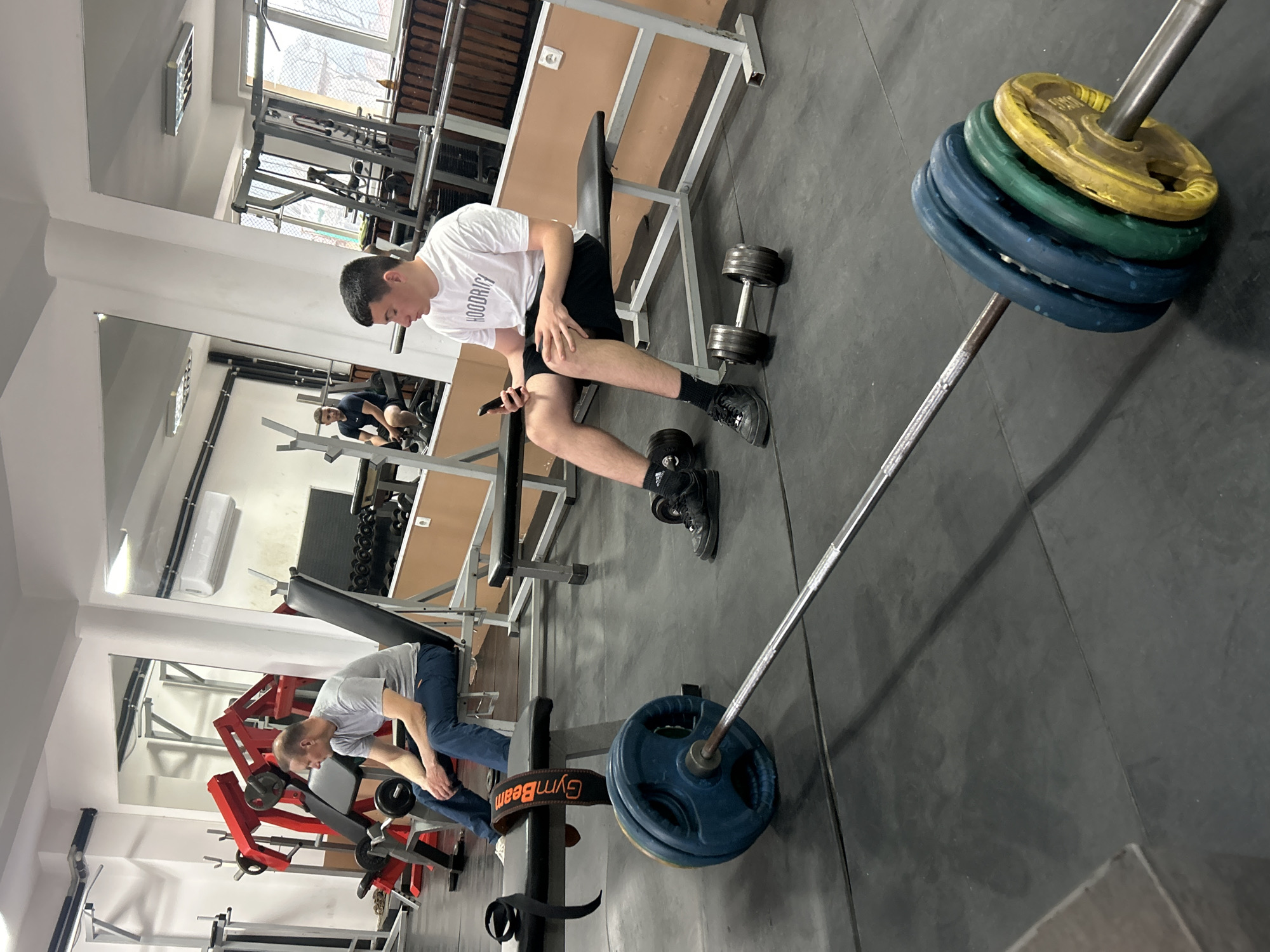 The image depicts an indoor gym setting in Zlatograd, Bulgaria, during daytime. The scene is captured with a slightly tilted perspective, focusing on the workout area.
In the mid-ground, a young man is seated on a black weight bench, engaged with his smartphone. He is wearing a white t-shirt with the text "HOODRICH" on it, black shorts, and black athletic shoes with socks. His right hand rests on his left knee while he holds the phone in his left hand. Next to him on the dark grey rubber floor is a dumbbell with black weight plates. A long barbell with colorful plates (blue, green, yellow) is prominently displayed, extending diagonally across the floor; a yellow plate is partially visible, showing "5 KG".
In the background, another man wearing a light grey t-shirt and blue jeans is bending over near a red and grey weight machine, appearing to adjust weights or equipment. A blue weight plate is visible near his feet, along with a "GymBeam" lifting belt on the floor.
The gym features light-colored walls, large mirrors reflecting parts of the interior and an additional individual, and a window allowing natural daylight to illuminate the space. Trees and building structures are visible outside the window. The ceiling has white panels, ventilation grilles, and an air conditioning unit mounted on the wall. The overall activity suggests a break or preparation during a workout session.