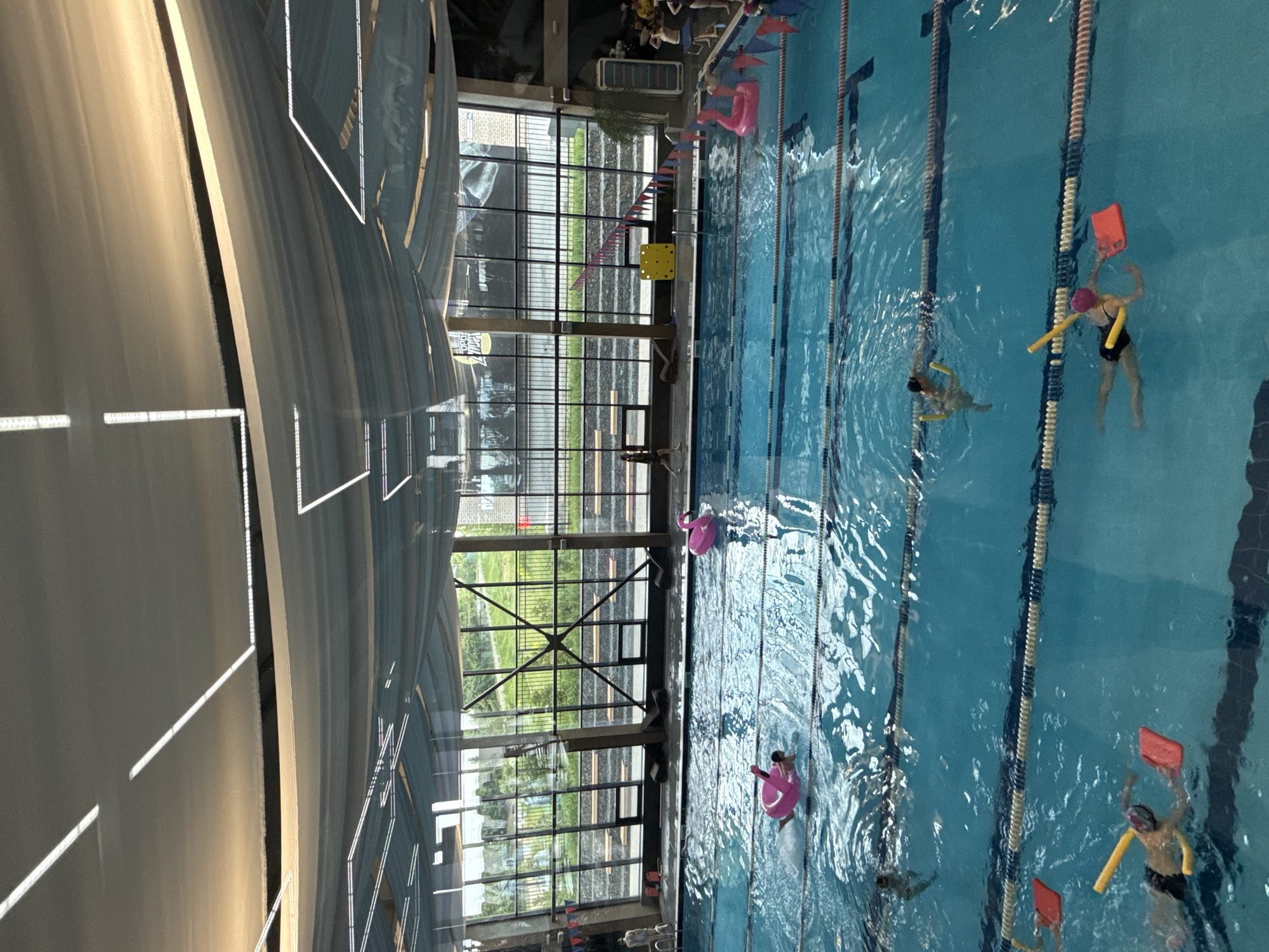 The image captures an indoor swimming pool facility during the daytime. The large, modern pool features multiple lanes marked by blue and white dividers. Several children are seen in the water, engaged in swimming activities, likely lessons or recreational play, utilizing various floatation devices.
In the foreground, a child wearing a black swimsuit and pink cap holds a yellow foam noodle and a red kickboard. Nearby, another child is partially visible with a noodle. Further along, a child in black swim trunks and goggles uses a yellow noodle and a red kickboard. Two other children are floating on distinctive pink inflatable flamingo rings. An adult figure is partially visible on the pool deck in the distance, near what appears to be starting blocks and some colorful flags.
The facility boasts a high, arched ceiling equipped with linear strip lighting, which casts bright reflections on the water and the ceiling's surface. A prominent feature is the expansive, floor-to-ceiling windows along one side, framed by dark structural beams. These windows offer a clear view of the outdoors, revealing a grassy area with fencing and distant buildings under what appears to be a bright, clear sky. A yellow square object sits on the pool deck near the windows. The scene suggests a vibrant and functional aquatic center, possibly in Votkinsk, Russia. No discernible text is visible.